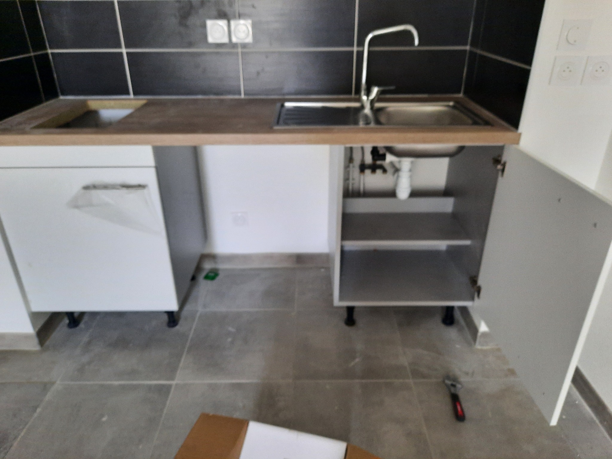 Częściowo zamontowany zlew kuchenny z otwartą szafką, widoczne rury i klucz francuski na podłodze. Czarno-białe płytki na ścianie w tle.