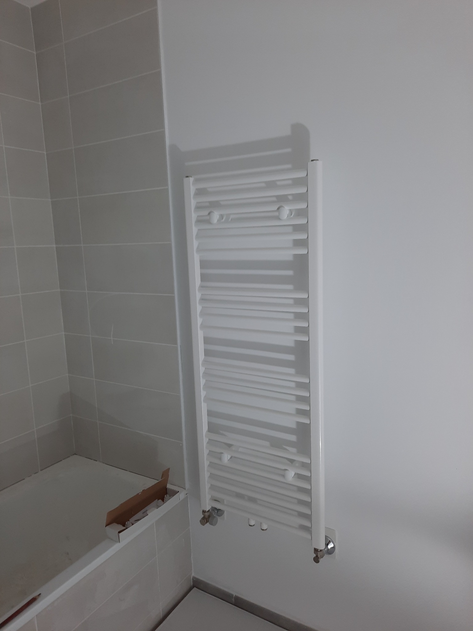 Biały, drabinkowy grzejnik łazienkowy zamontowany na ścianie w jasnej łazience z szarymi płytkami. Widoczna wanna i karton z akcesoriami.