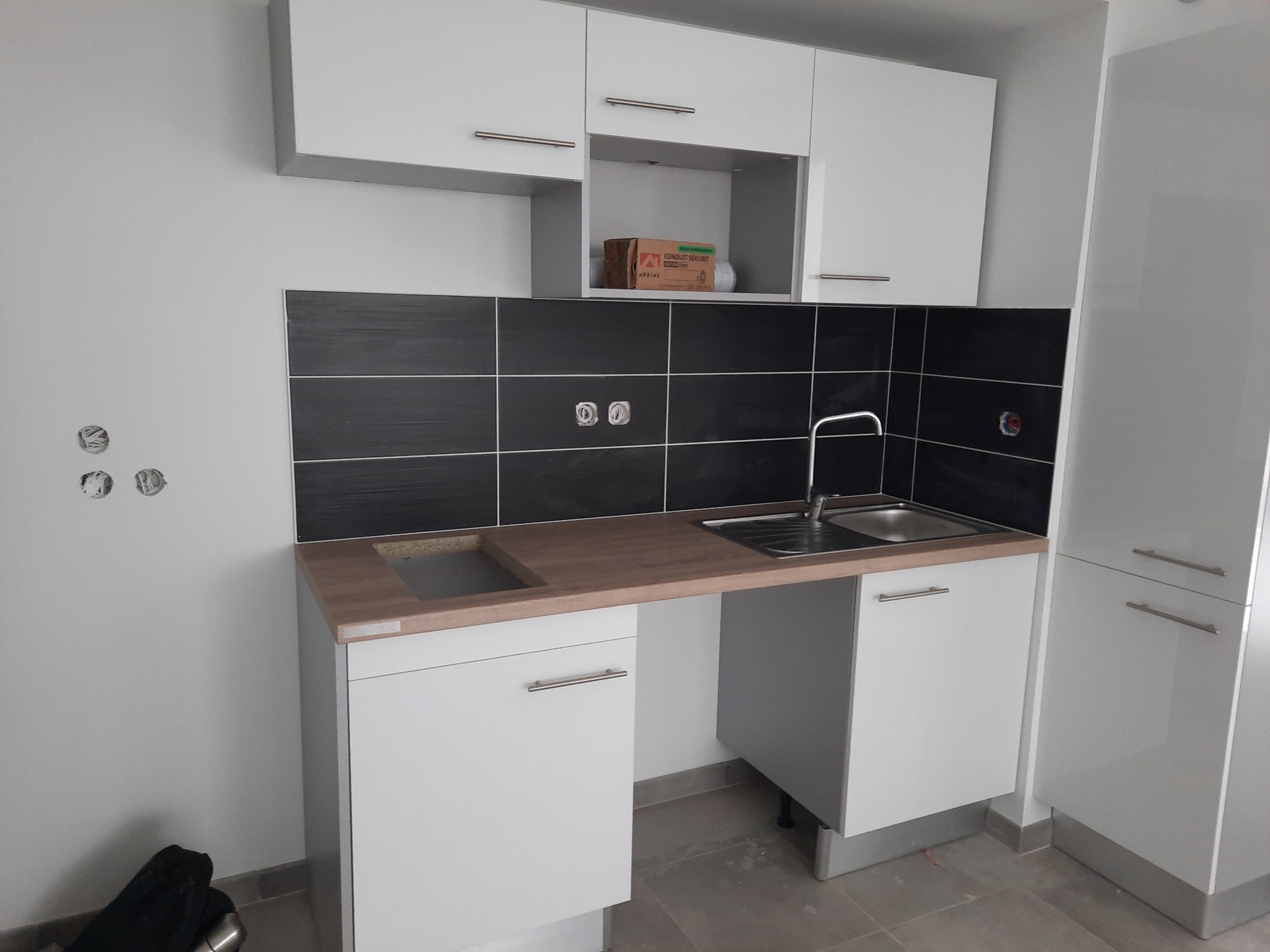 Nowoczesna, biała zabudowa kuchenna z drewnianym blatem i czarnymi płytkami. Widoczny zlew i otwór na płytę indukcyjną. W tle niezabudowane gniazdka elektryczne.