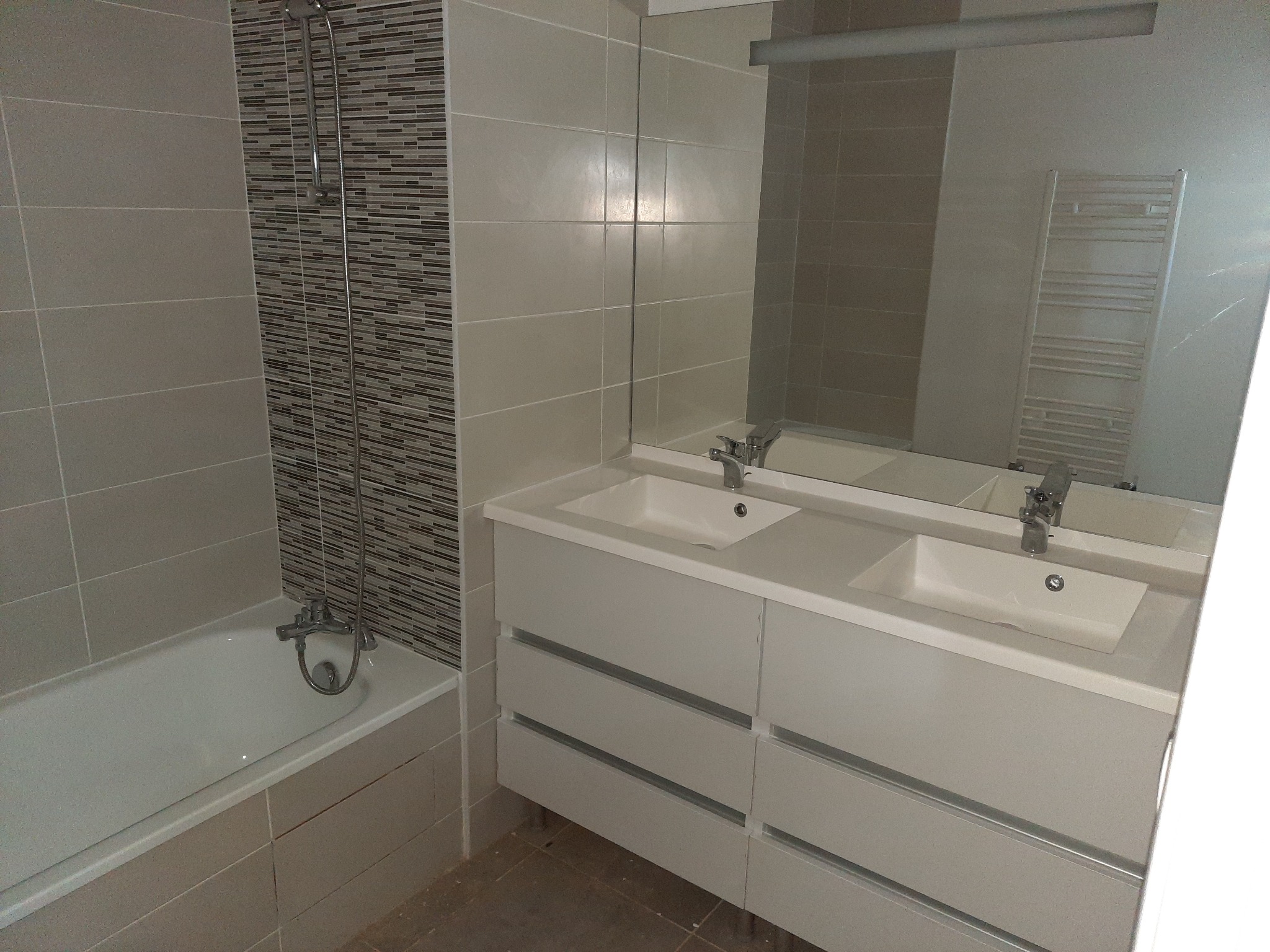 Nowoczesna łazienka w neutralnych kolorach: wanna z baterią, umywalki w zabudowie z lustrem i grzejnik drabinkowy. Minimalistyczny design, płytki w odcieniach beżu i brązu.