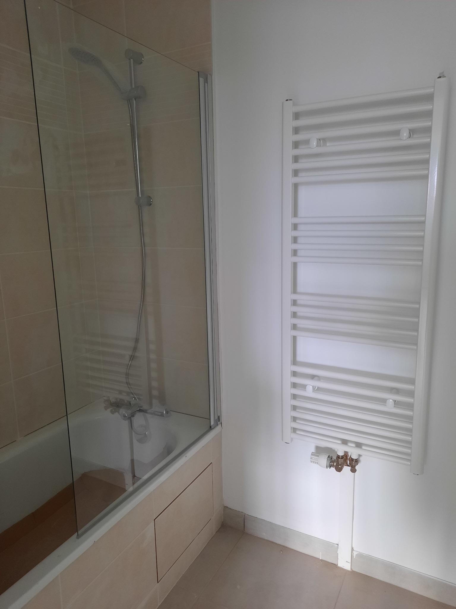 Biały grzejnik łazienkowy zamontowany na ścianie obok wanny z parawanem i baterią. Widoczne podłączenia hydrauliczne grzejnika.