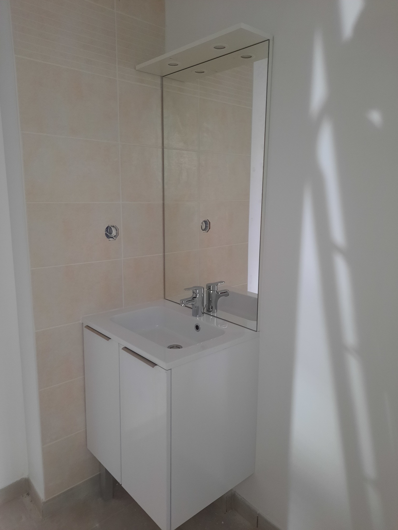 Biała szafka z umywalką i lustrem z oświetleniem w łazience z beżowymi płytkami. Widoczny kran i fragment ściany. Minimalistyczny design.