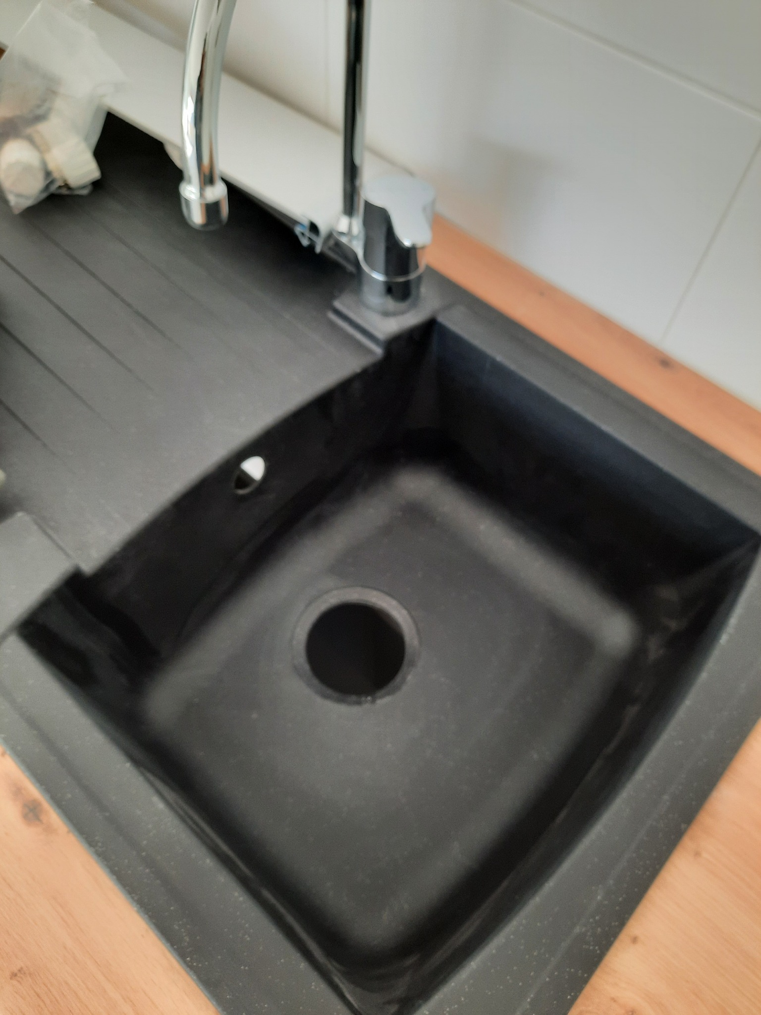Czarny, granitowy zlew kuchenny z chromowaną baterią, zamontowany w blacie. Widoczny fragment syfonu i akcesoria montażowe. Górny kadr.
