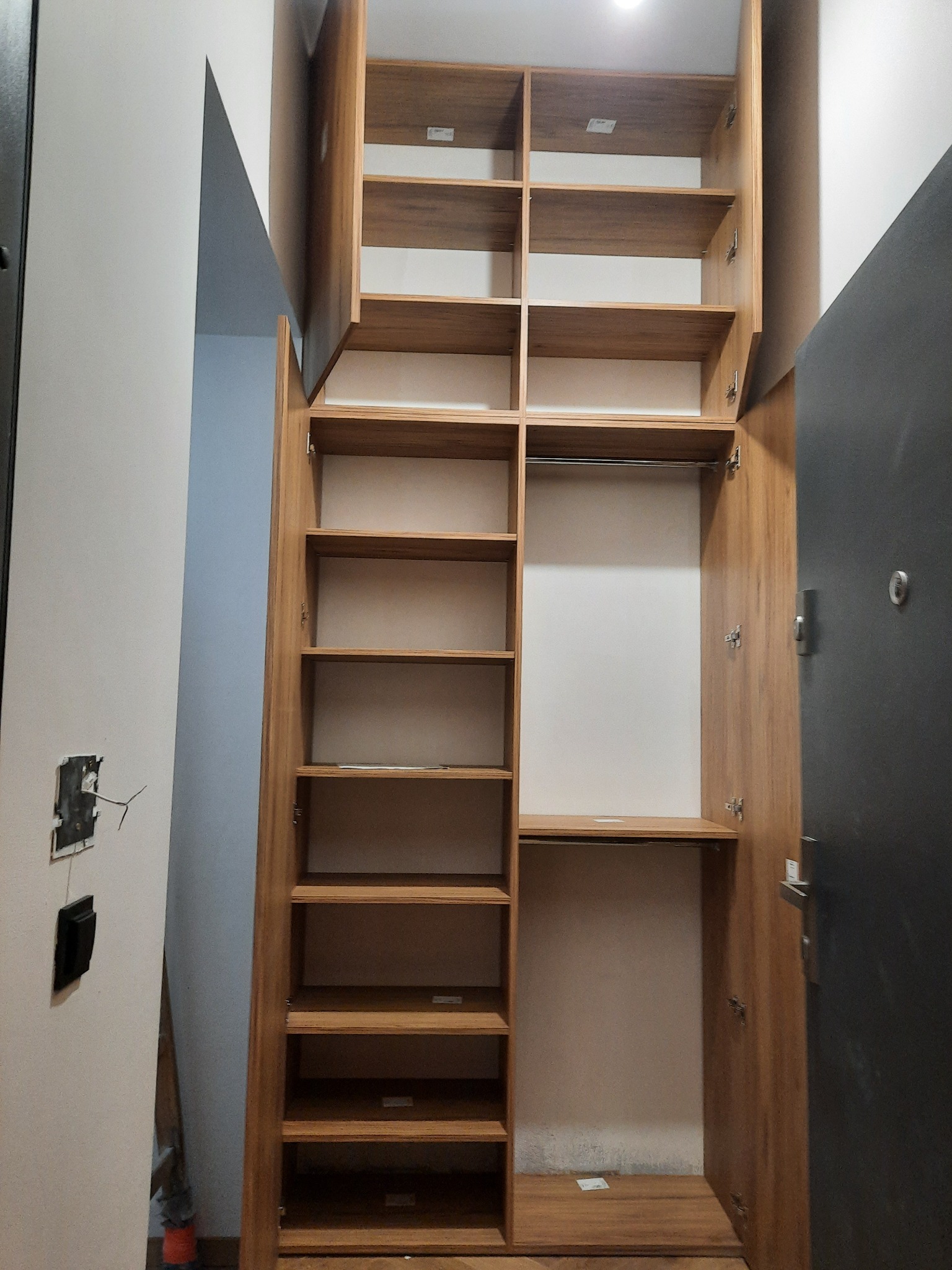 Wysoka, drewniana szafa wnękowa z otwartymi drzwiami, ukazująca półki i drążek na ubrania. Widoczny fragment ściany z wystającymi przewodami elektrycznymi.