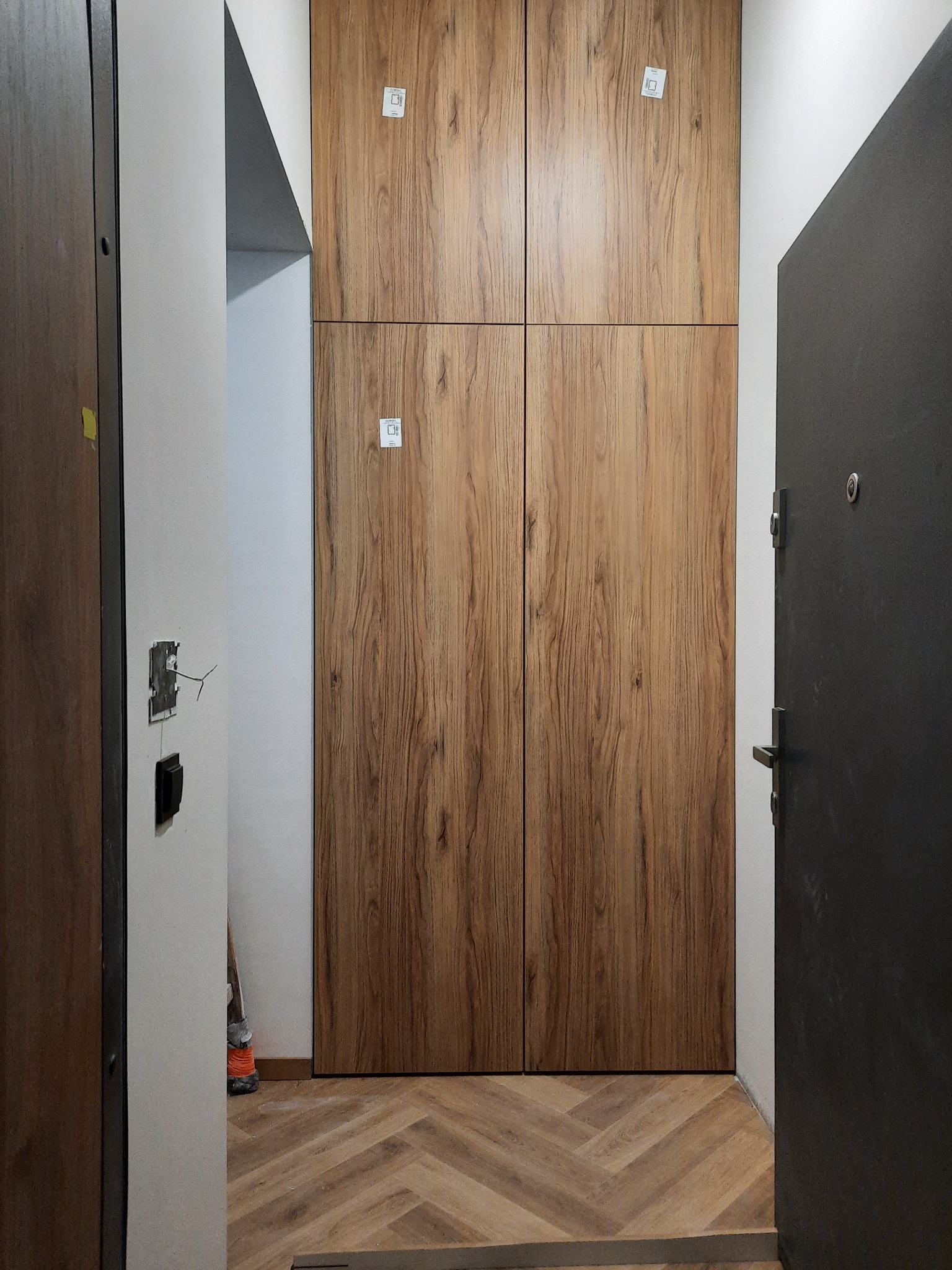 Szafa wnękowa z drewnianymi frontami, zamontowana w korytarzu z parkietem ułożonym w jodełkę. Widoczna instalacja elektryczna w ścianie.