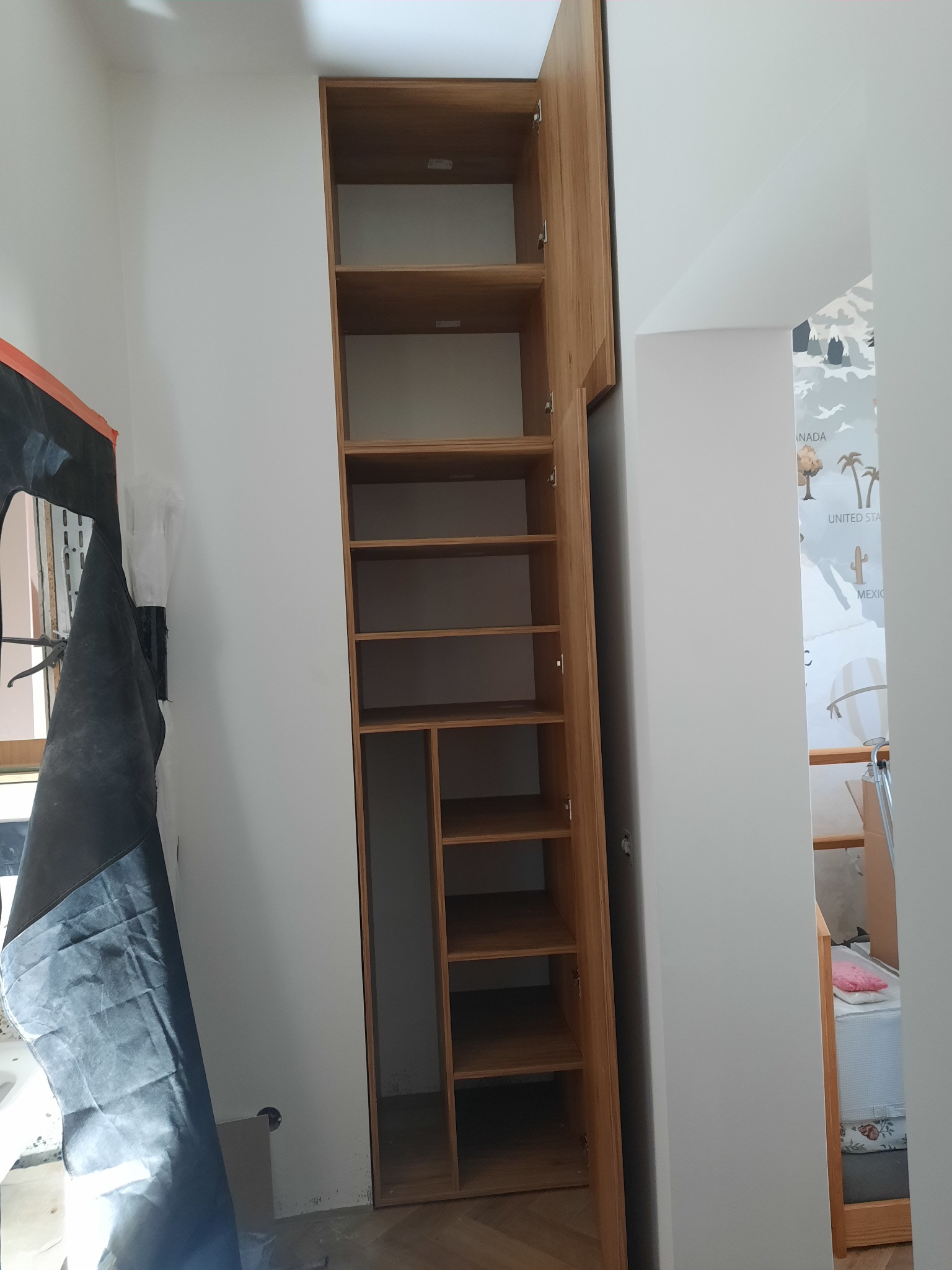Wysoka, drewniana szafa wnękowa z otwartymi półkami, zamontowana w rogu pokoju, obok okna zasłoniętego folią malarską. Widoczne elementy wykończeniowe.