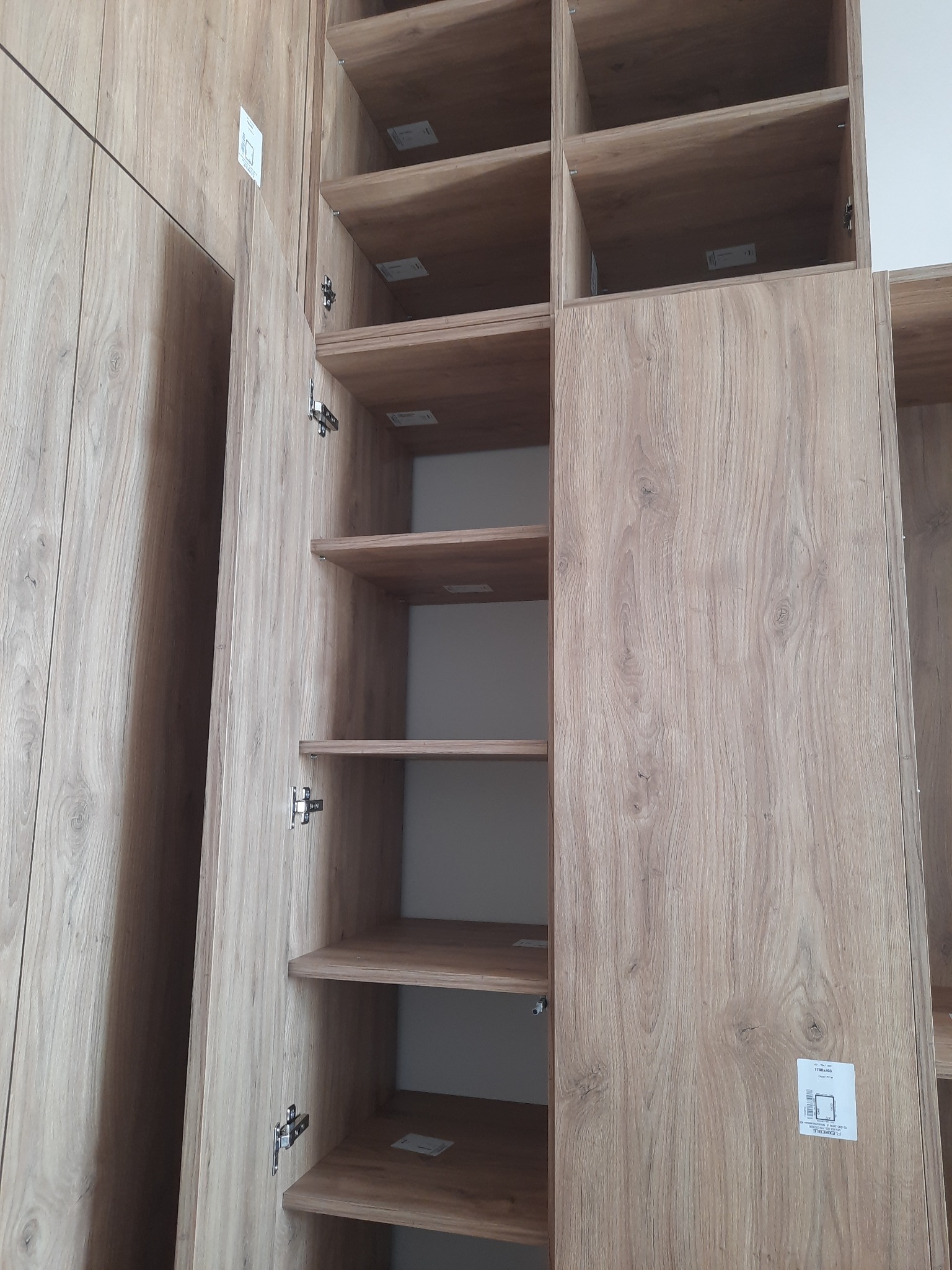 Zamontowana szafa z otwartymi drzwiami i półkami o fakturze drewna. Widoczne zawiasy i etykiety na wewnętrznych ścianach. Ujęcie z bliska, w neutralnym oświetleniu.