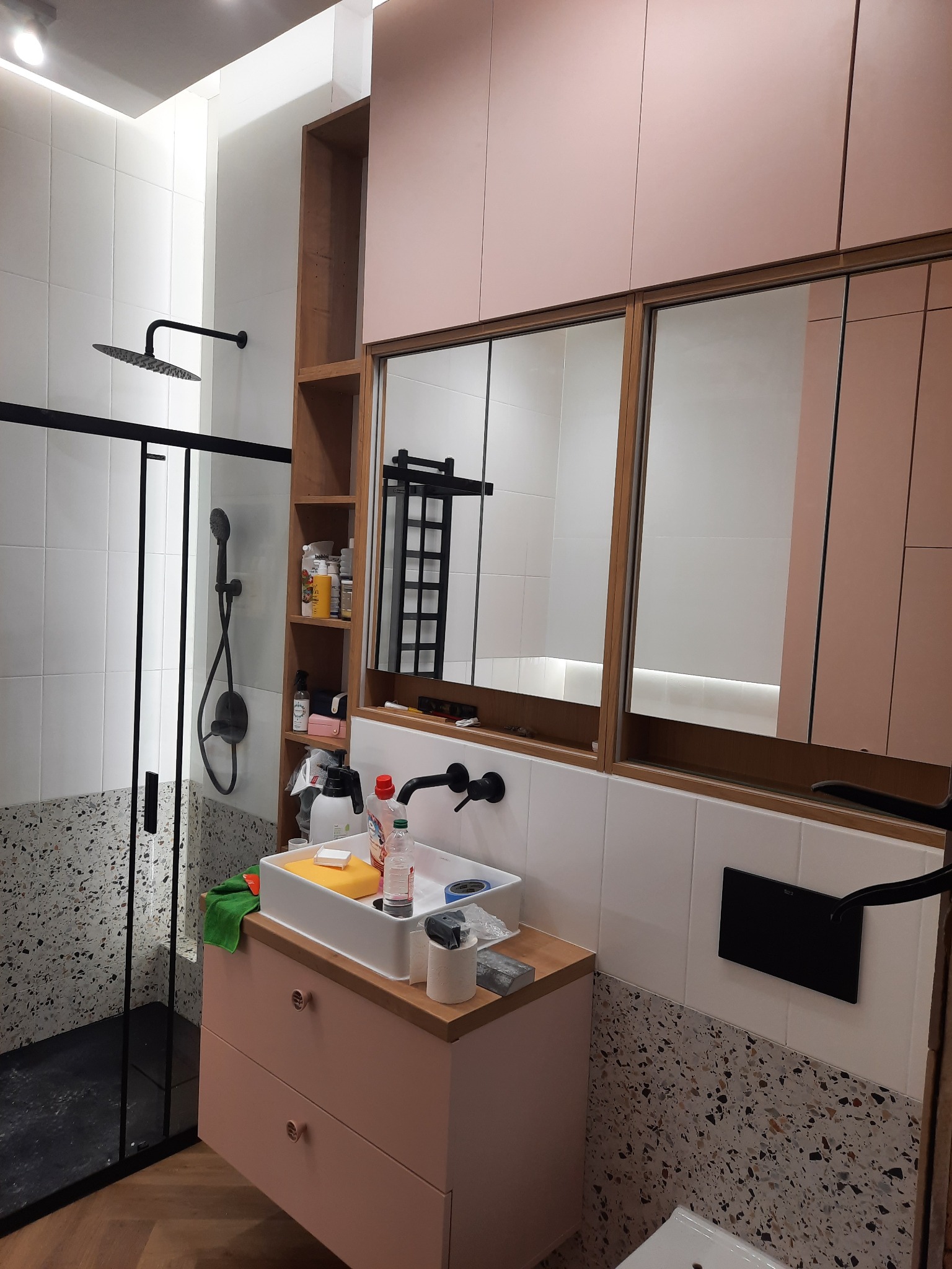 Nowoczesna łazienka z kabiną prysznicową typu walk-in, szafką umywalkową w odcieniach różu i czarną armaturą. Widoczne półki z kosmetykami i lustra.