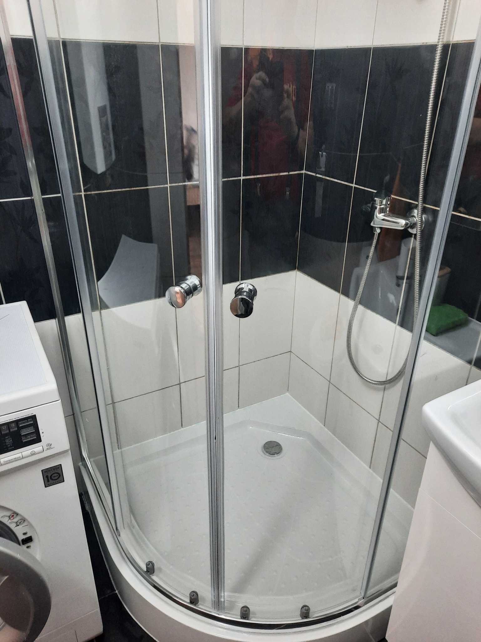 Zamknięta kabina prysznicowa z chromowanymi uchwytami, przylegająca do pralki i umywalki na tle czarno-białych płytek w łazience.