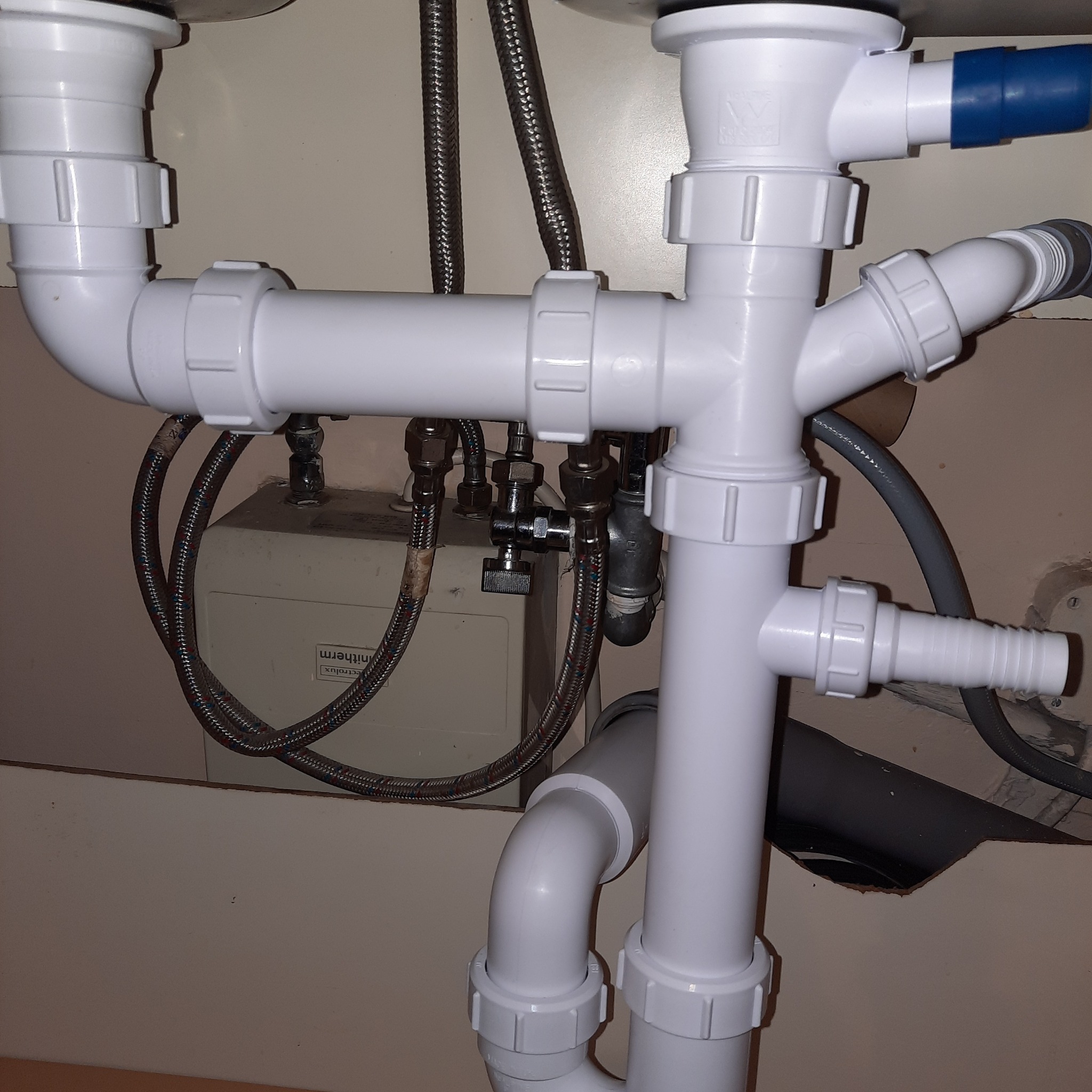 Złożony system białych rur odpływowych pod zlewem, z widocznymi elementami mocującymi i metalowymi wężykami, zainstalowany w szafce z kremowym tłem.