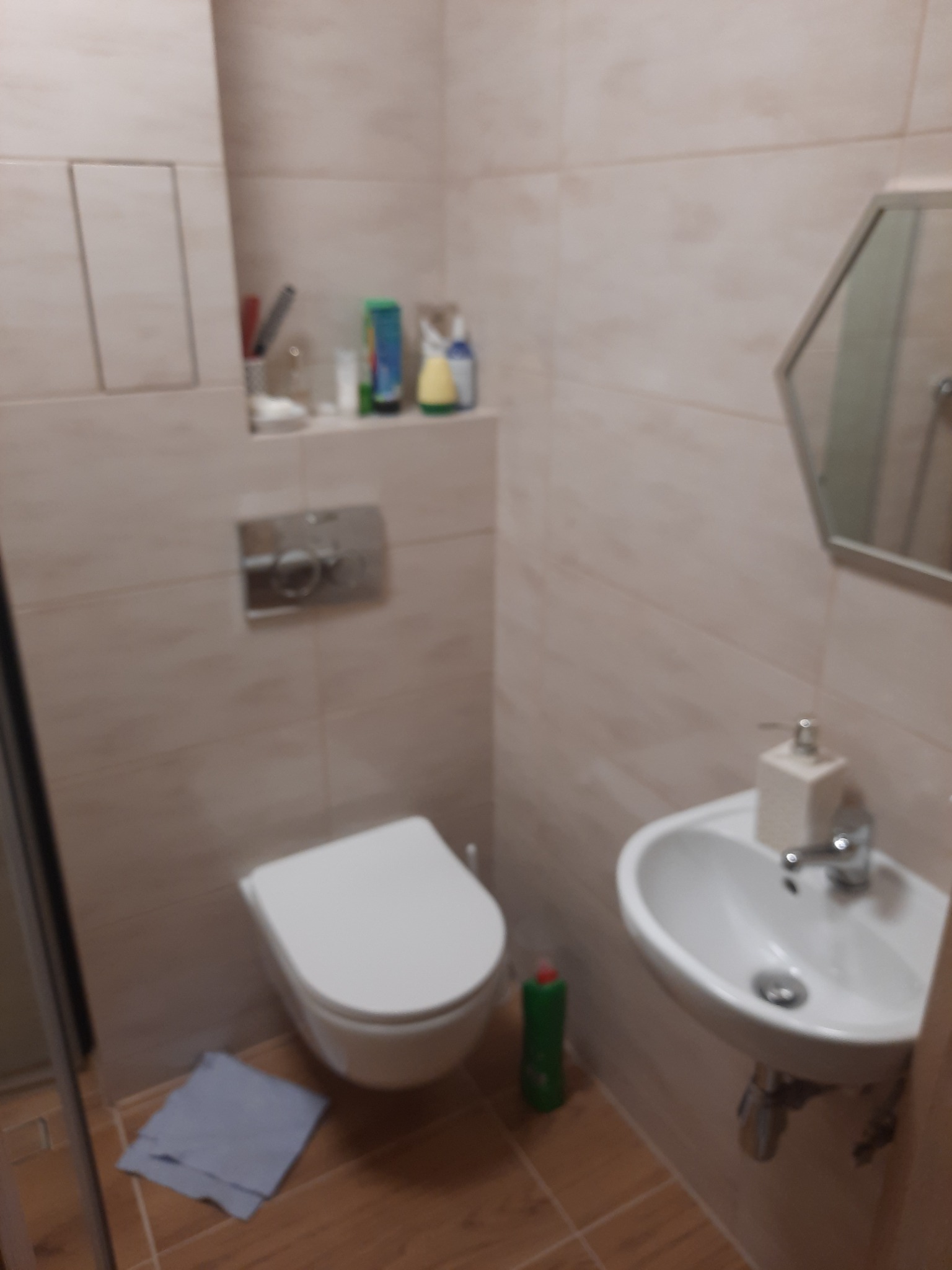 Jasna łazienka z umywalką, toaletą podwieszaną i heksagonalnym lustrem. Płytki imitujące drewno na podłodze i ścianach. Widoczny fragment kabiny prysznicowej.