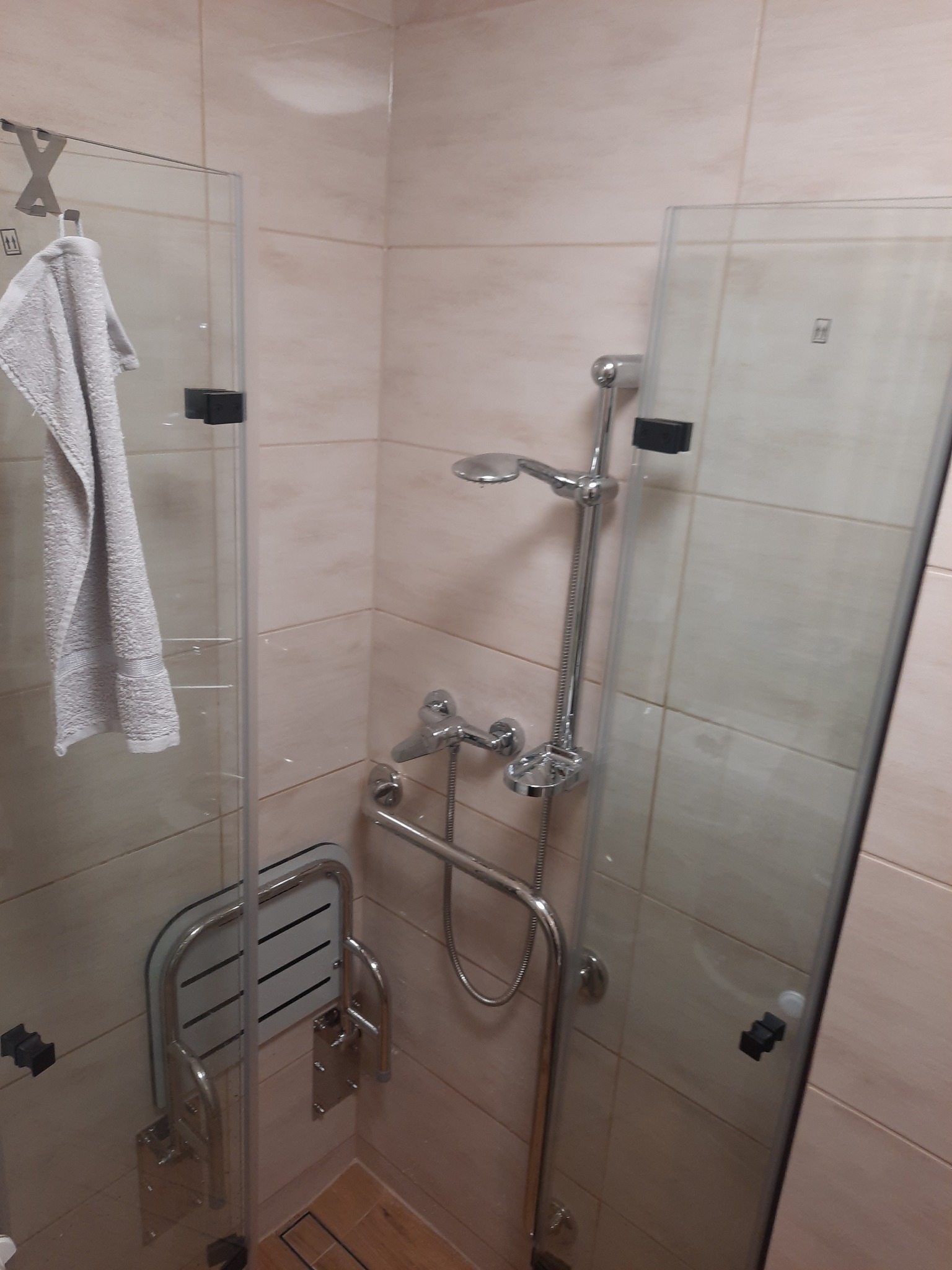 Kabina prysznicowa z siedziskiem i uchwytami dla niepełnosprawnych, widok z boku, beżowe płytki, chromowana armatura. Na szybie wisi ręcznik.