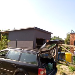 Valdibud - Nowoczesny, szary budynek gospodarczy z płaskim dachem, widok zza otwartego bagażnika samochodu kombi, w tle ceglany dom i ogrodzenie.