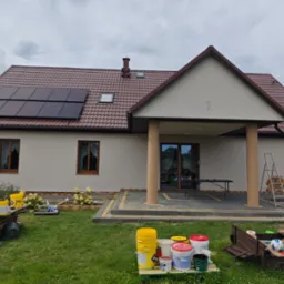 Jednopiętrowy dom z brązowym dachem, panelami słonecznymi, pomalowaną elewacją w odcieniach beżu, widoczne wiadra z farbą i drabina, sugerujące prace wykończeniowe na zewnątrz.