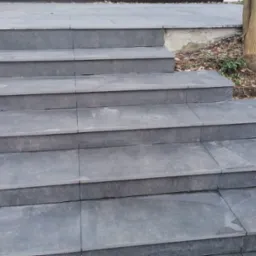 Zewnętrzne schody z szarych płyt kamiennych prowadzące do budynku, otoczone korą drzewną i roślinnością.