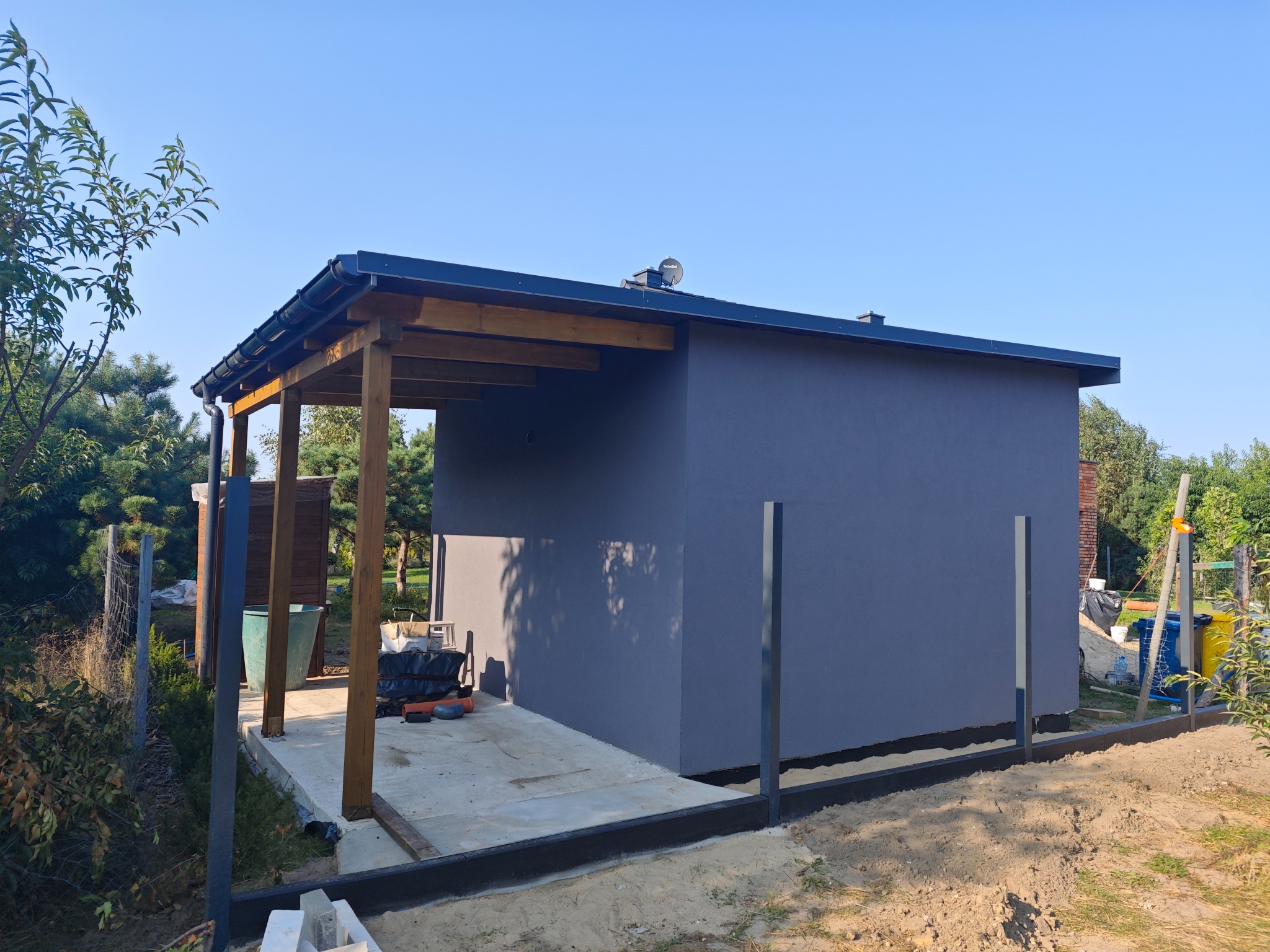 Nowoczesny, szary budynek gospodarczy z drewnianą konstrukcją zadaszenia, widoczny w trakcie budowy ogrodzenia na piaszczystym terenie.