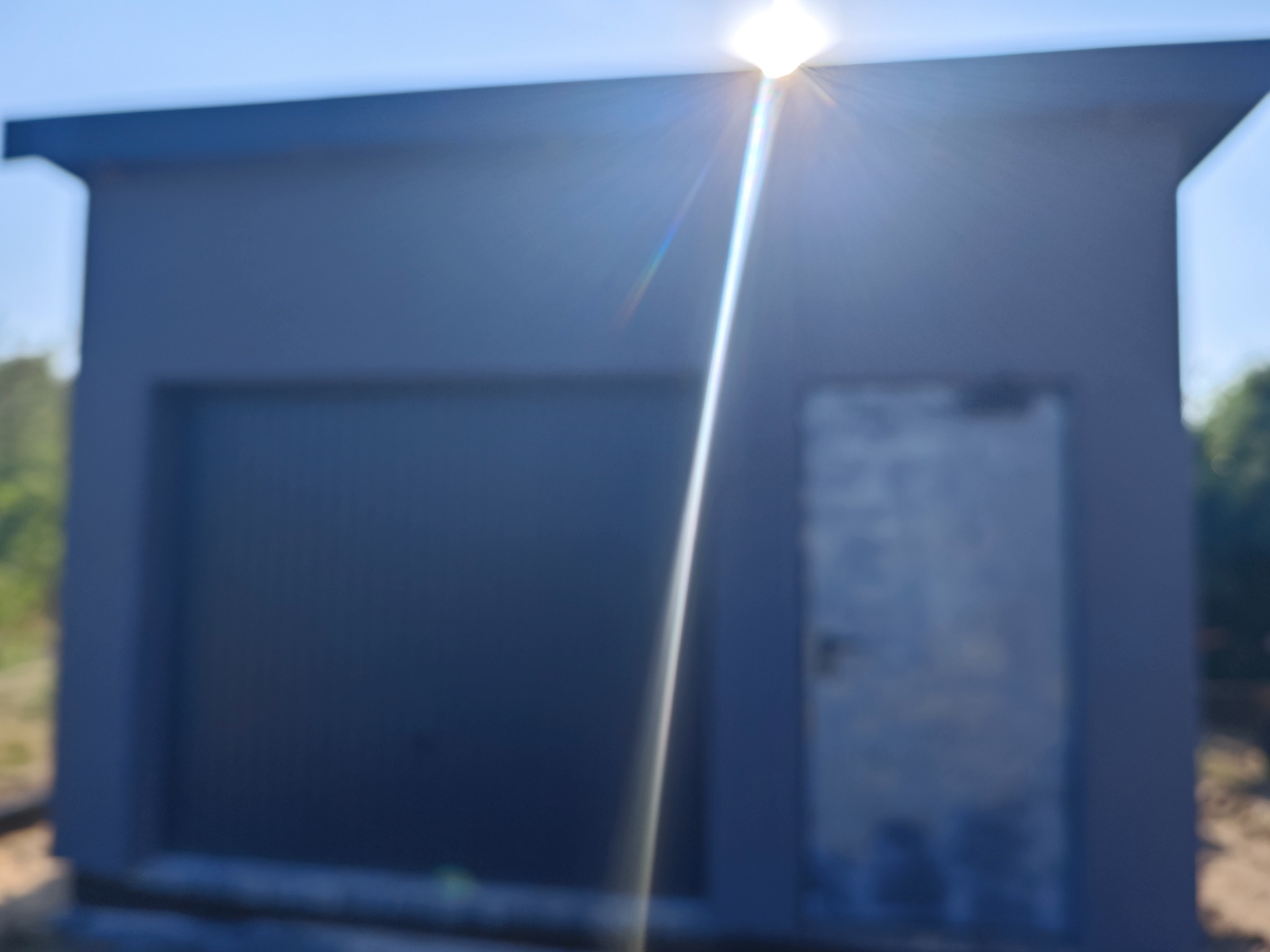 Nowo pomalowany, szary garaż z bramą segmentową i drzwiami wejściowymi, uchwycony w silnym świetle słonecznym, powodującym odblaski.