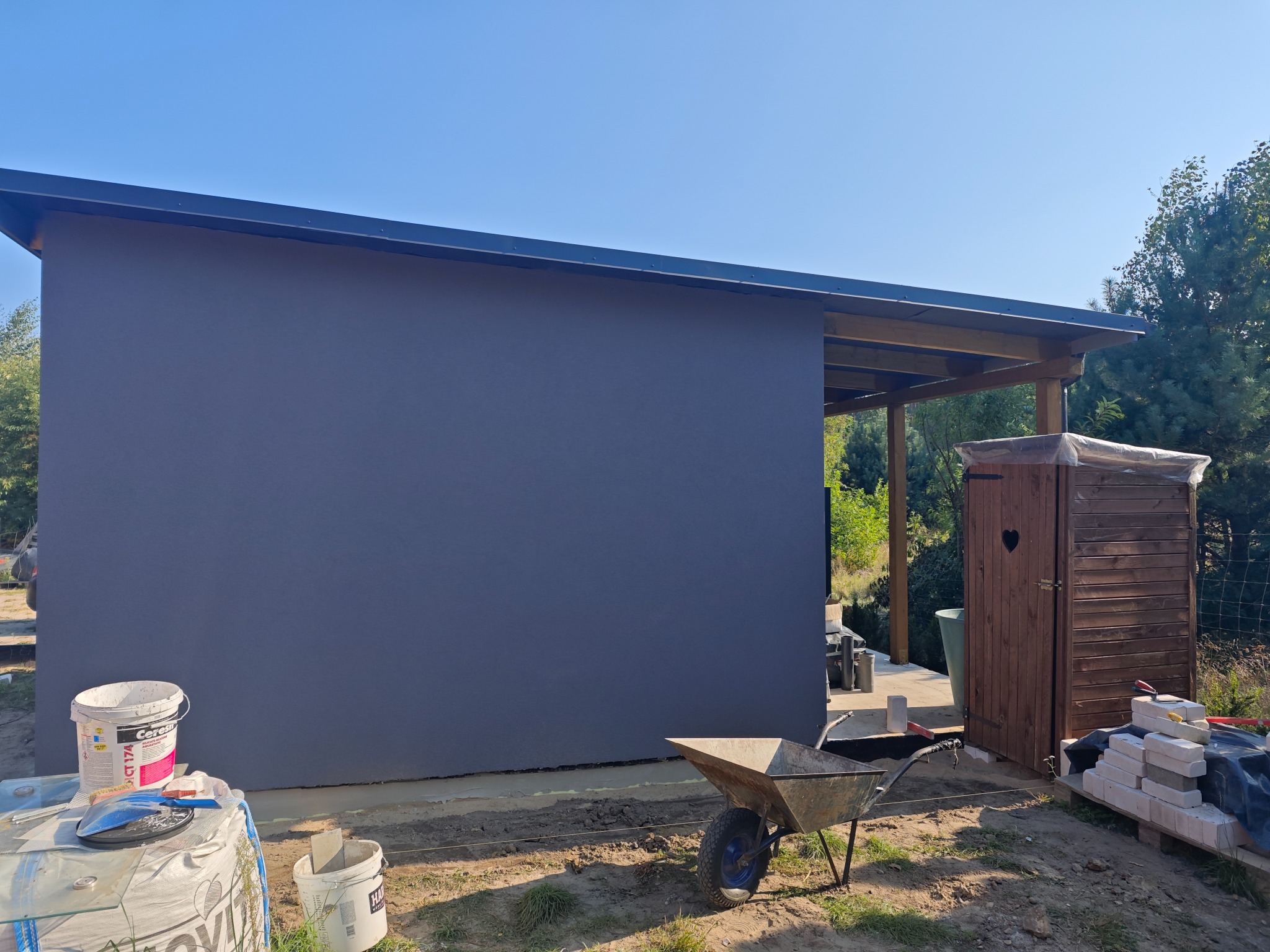 Świeżo pomalowana, ciemnoszara ściana budynku gospodarczego z widocznymi wiadrami z farbą, taczką i drewnianą toaletą z serduszkiem w drzwiach, w słoneczny dzień.