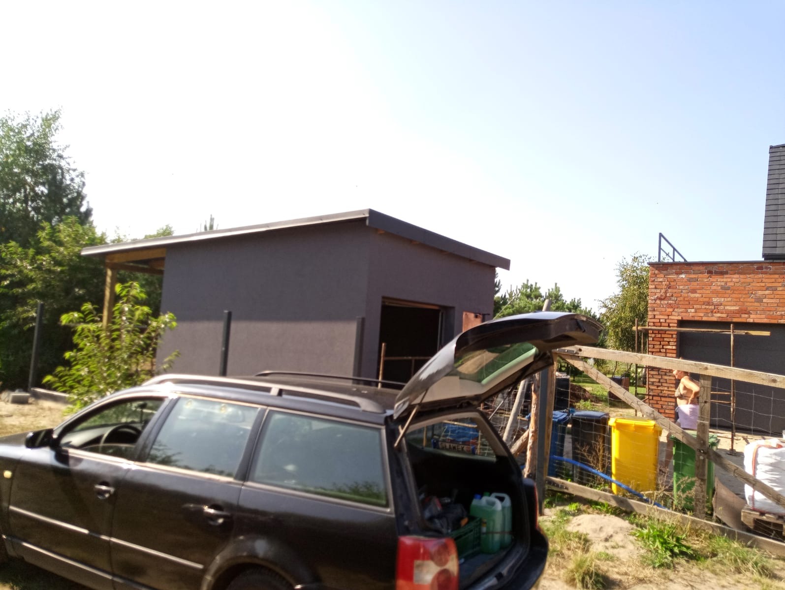 Nowoczesny, szary budynek gospodarczy z płaskim dachem, widok zza otwartego bagażnika samochodu kombi, w tle ceglany dom i ogrodzenie.