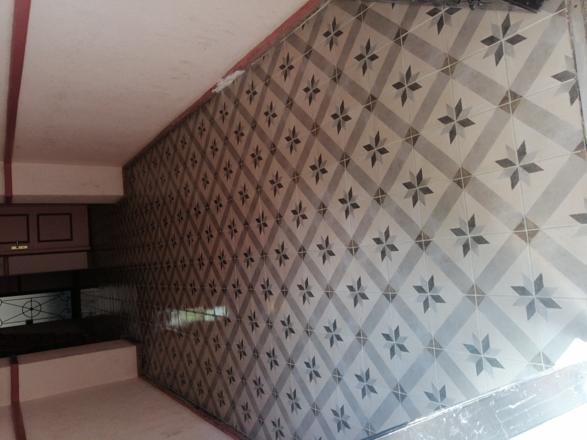 Ściana i podłoga wyłożone szaro-białymi płytkami ceramicznymi w geometryczny wzór gwiazd, widok z dołu, fragment szafki oraz okna w tle.