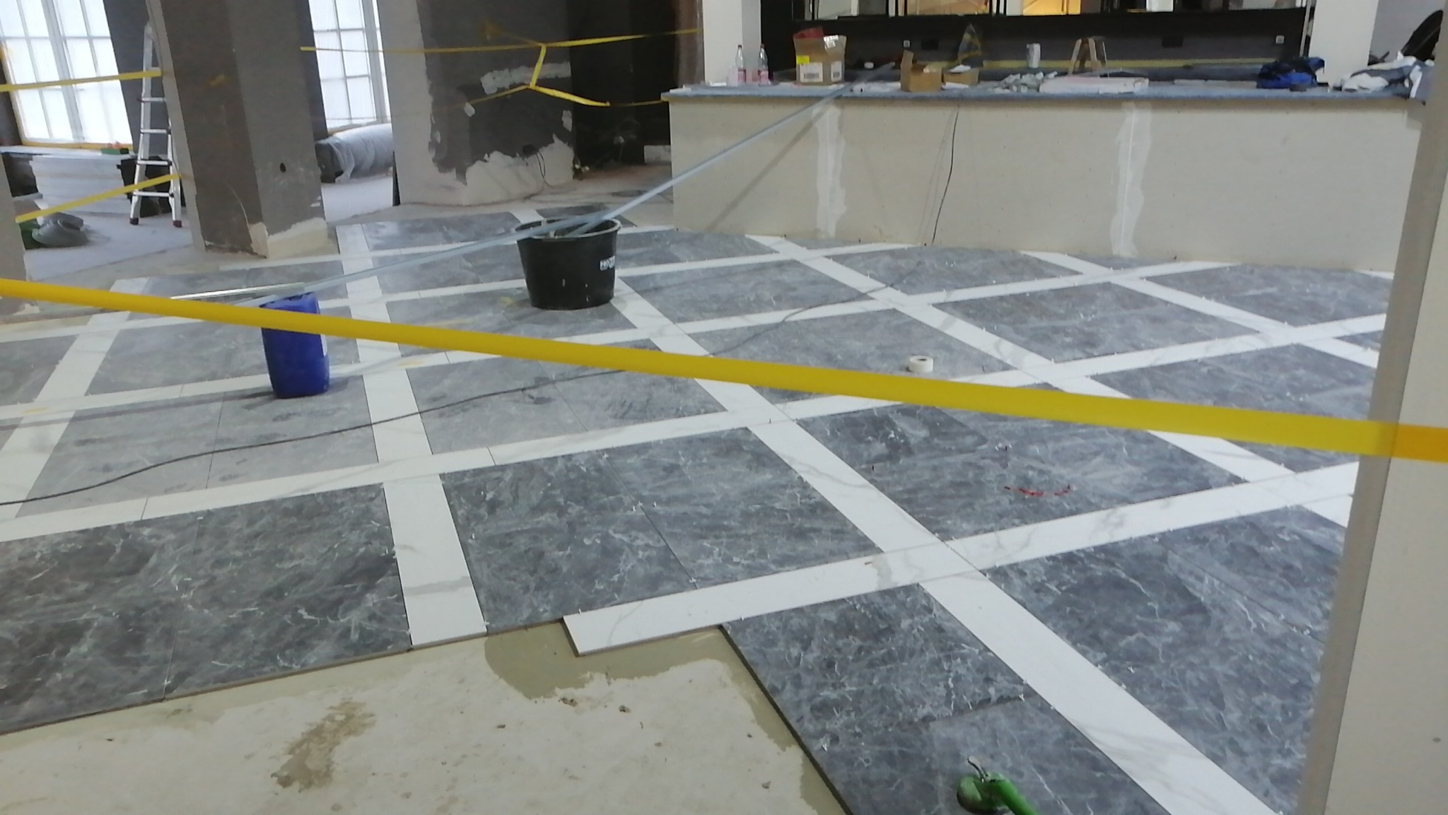 Układanie marmurowej podłogi w kwadraty z białymi wstawkami, widoczny kubeł, niebieski pojemnik i taśma ostrzegawcza.
