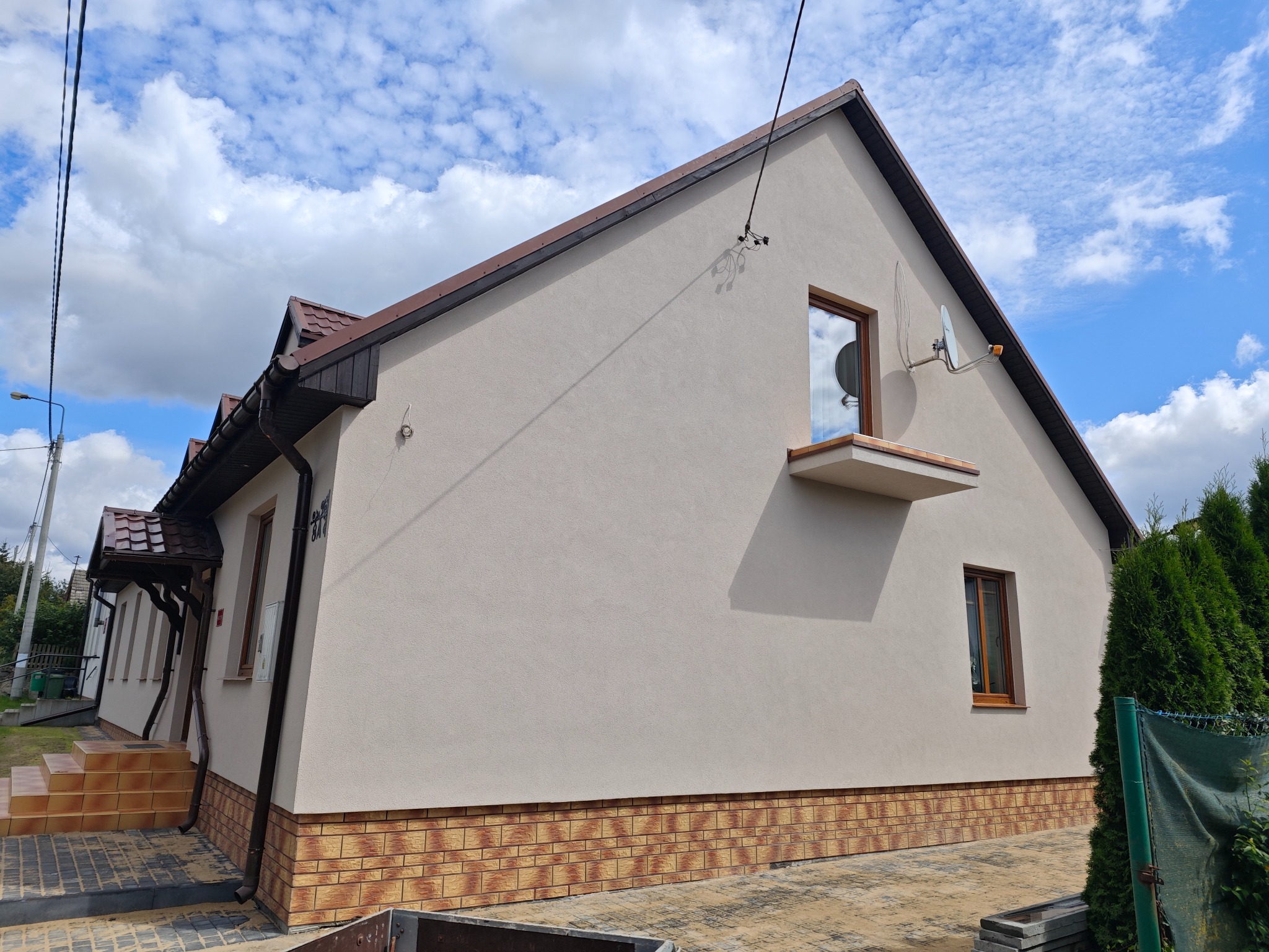 Elewacja domu jednorodzinnego po remoncie, z widocznym ociepleniem i nową strukturą tynku, brązowe okna i dachówka, antena satelitarna na ścianie szczytowej.