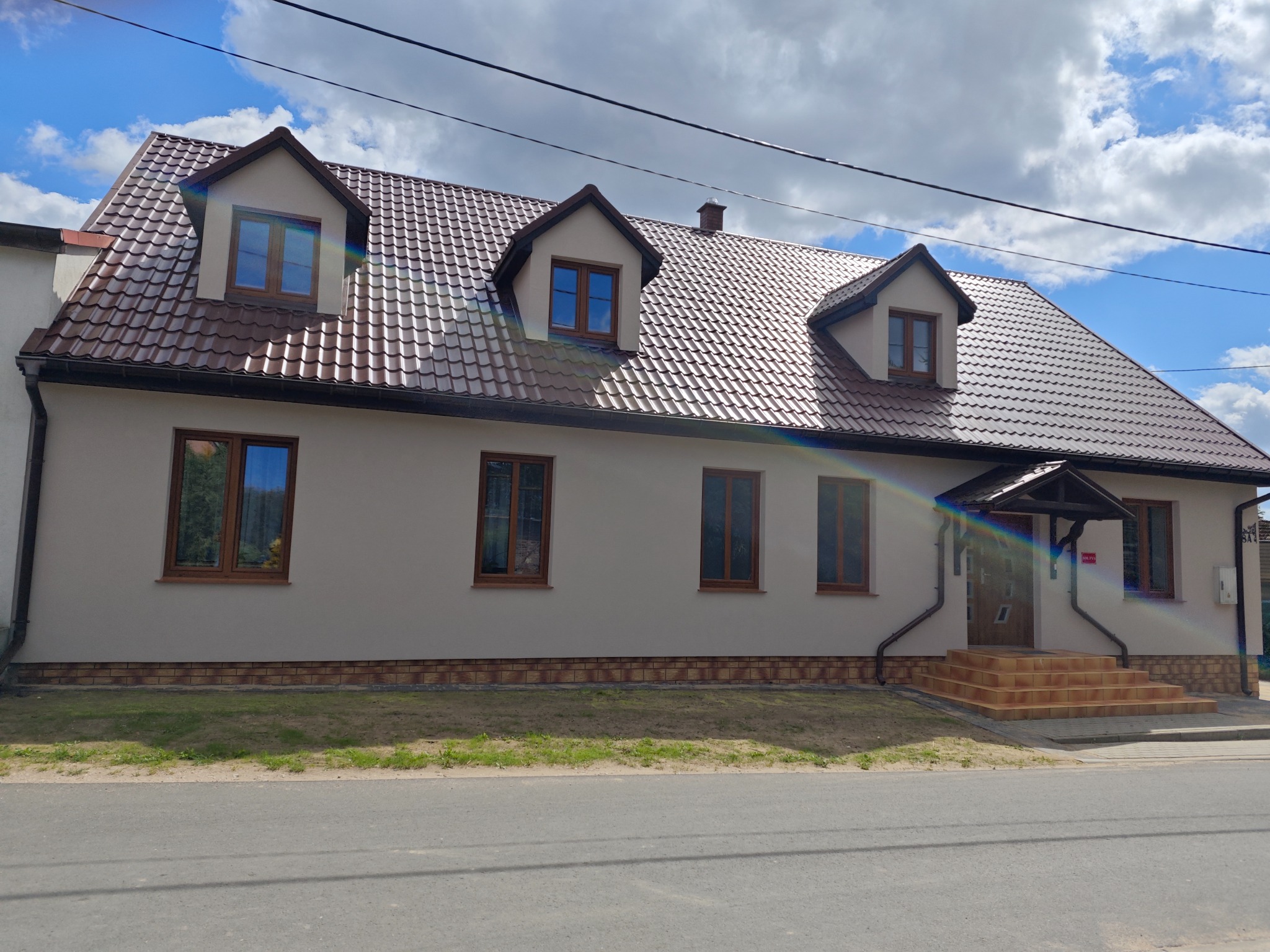 Budynek jednorodzinny z brązowym dachem i jasną elewacją, widoczne okna w brązowych ramach i wejście z kilkoma stopniami, na elewacji widoczny tęczowy efekt świetlny.