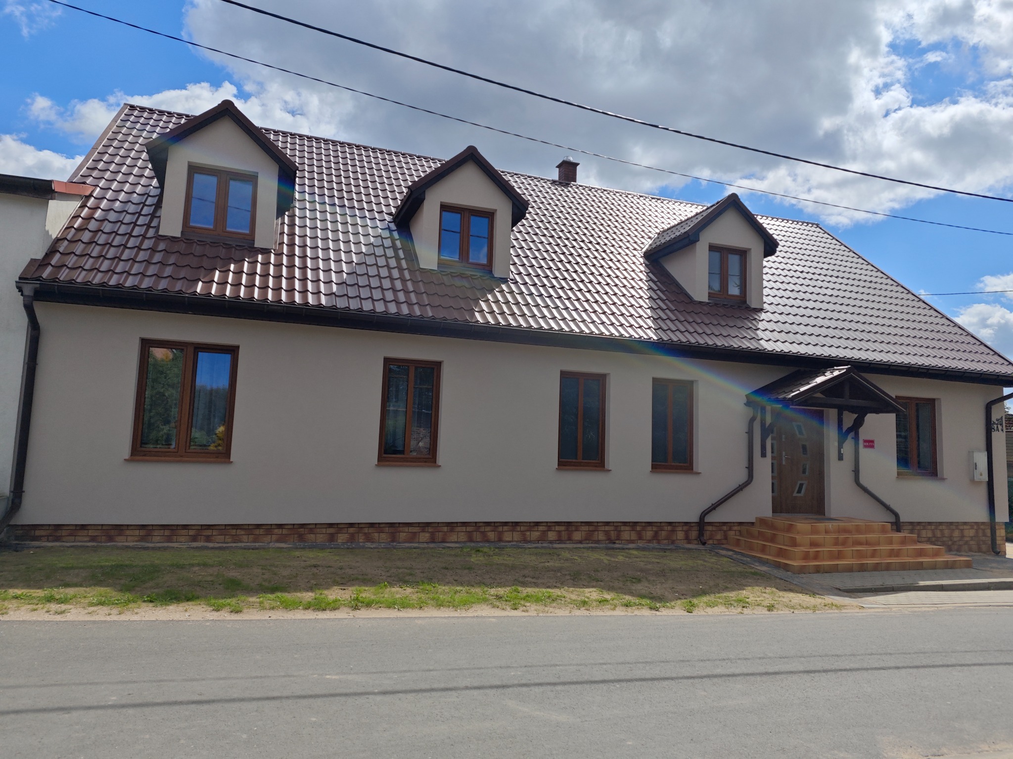 Jasny dom jednorodzinny z brązowym dachem i oknami, widoczny z poziomu ulicy w słoneczny dzień.