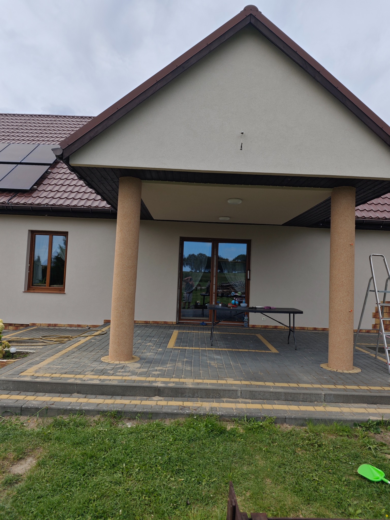 Dom jednorodzinny z jasną elewacją i brązowym dachem, widoczne panele solarne, kolumny wejściowe wykończone tynkiem mozaikowym, na podjeździe stół roboczy i drabina.