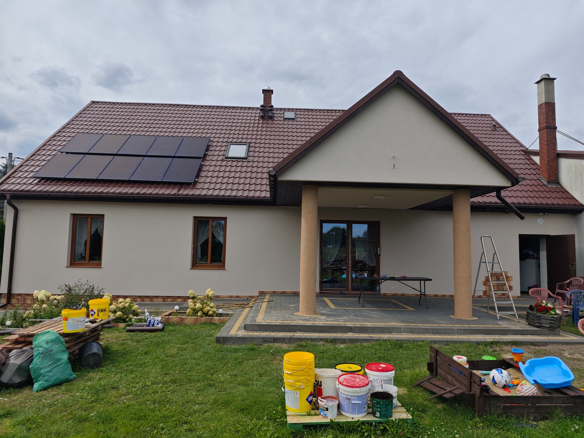 Jednopiętrowy dom z brązowym dachem, panelami słonecznymi, pomalowaną elewacją w odcieniach beżu, widoczne wiadra z farbą i drabina, sugerujące prace wykończeniowe na zewnątrz.