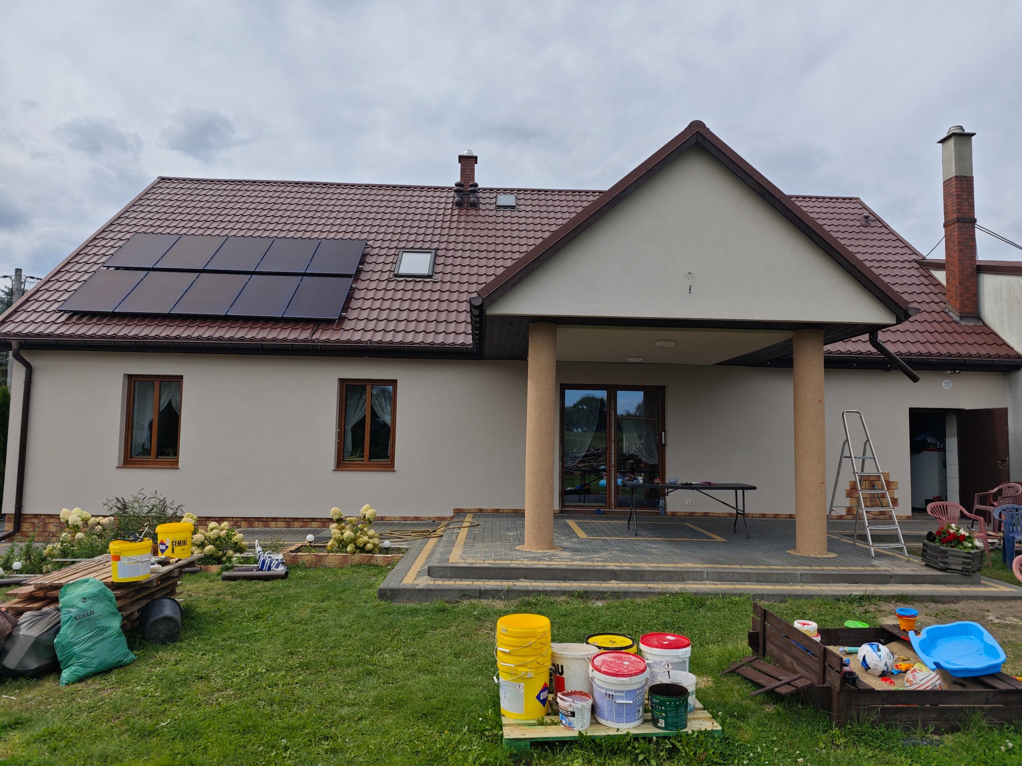 Ocieplony dom jednorodzinny z panelami słonecznymi na dachu, widoczne wiadra z farbą i materiały budowlane na trawniku przed elewacją.
