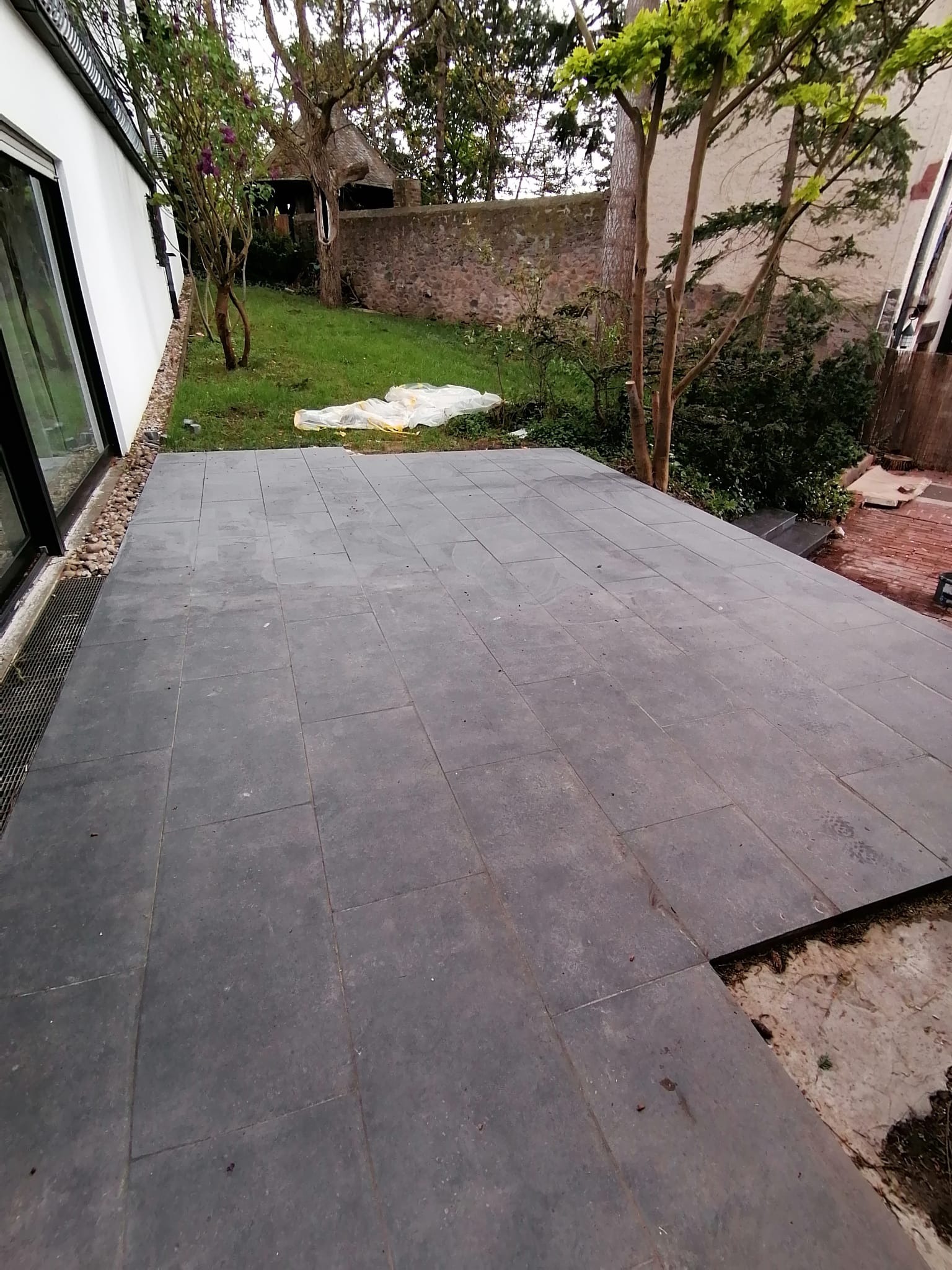 Szara, kwadratowa nawierzchnia tarasu z płyt kamiennych, częściowo ułożona, z widocznym fragmentem podłoża i ogrodem w tle.