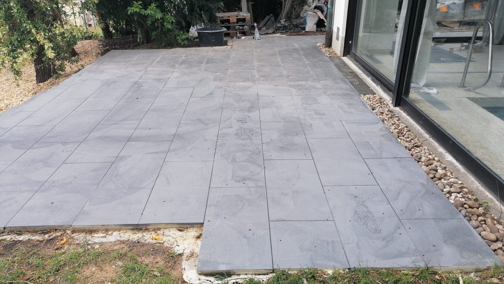 Układanie szarych płyt tarasowych na zewnątrz budynku, widoczne ślady stóp na powierzchni, obok okna z kamieniami ozdobnymi, paleta i wiadro w tle.