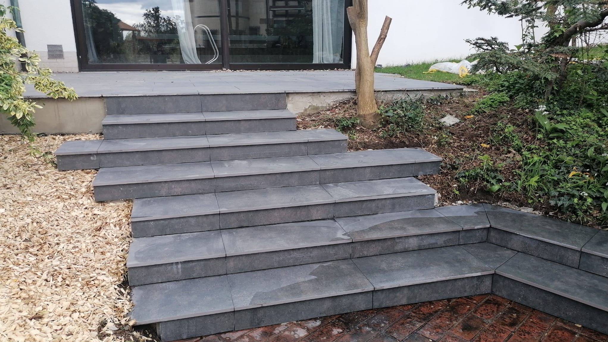Zewnętrzne schody z szarych płyt prowadzące do domu, otoczone korą drzewną i roślinnością, widok z dołu.