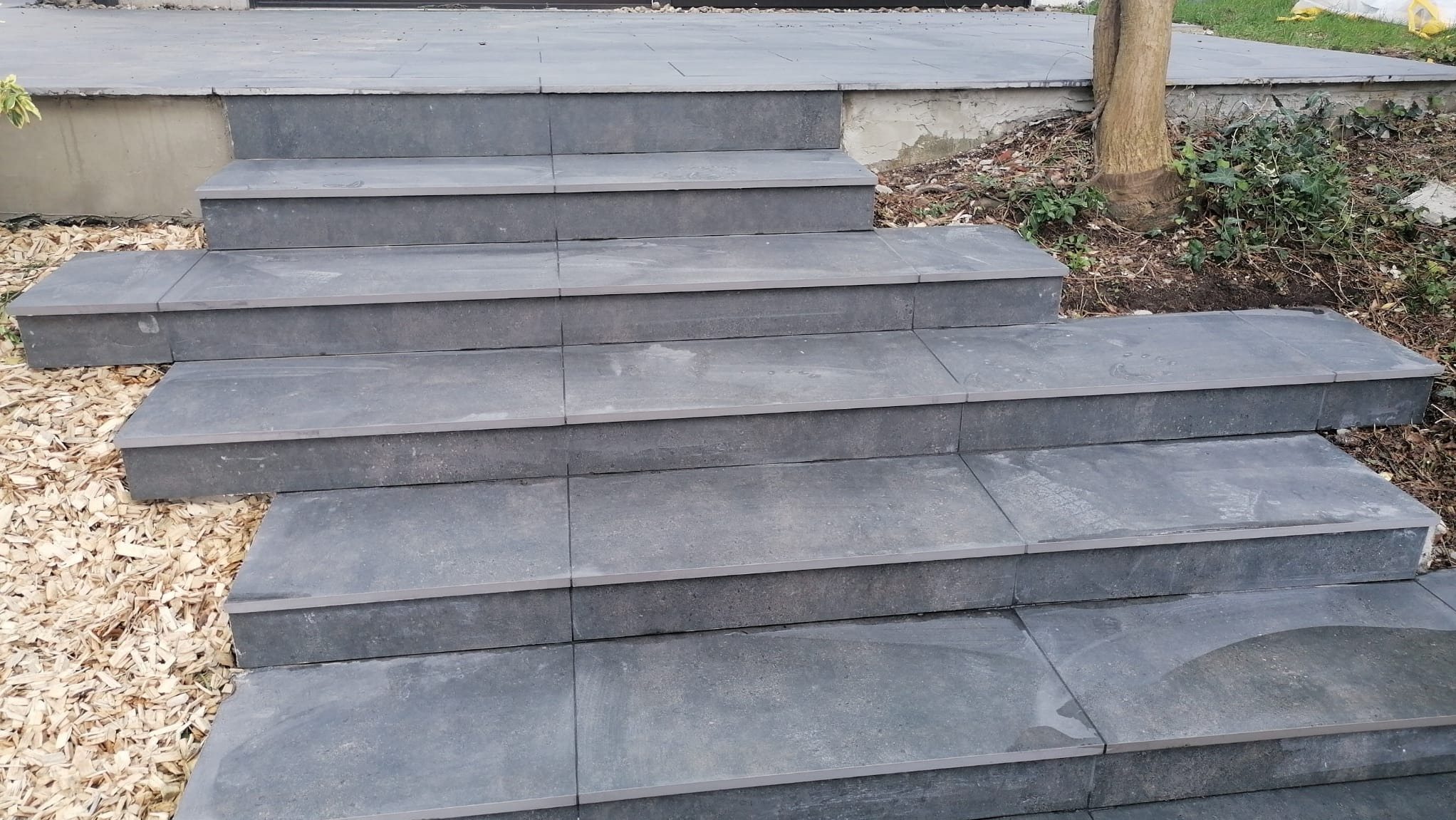 Zewnętrzne schody z szarych płyt kamiennych prowadzące do budynku, otoczone korą drzewną i roślinnością.