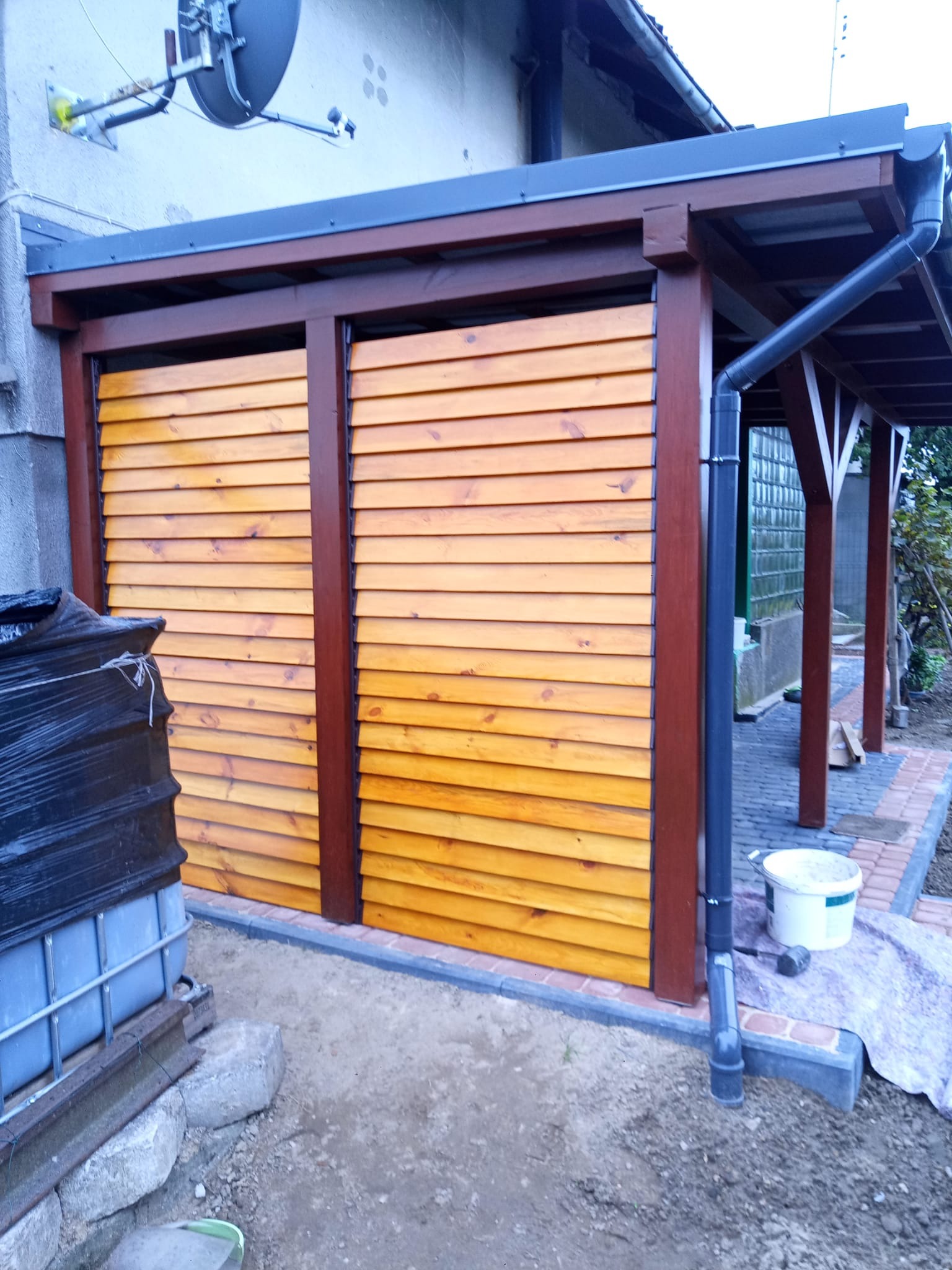 Zadaszenie tarasu z drewnianą konstrukcją i lamelami w kolorze miodowego drewna, widoczne rynny i kostka brukowa wokół, z boku budynku z anteną satelitarną.