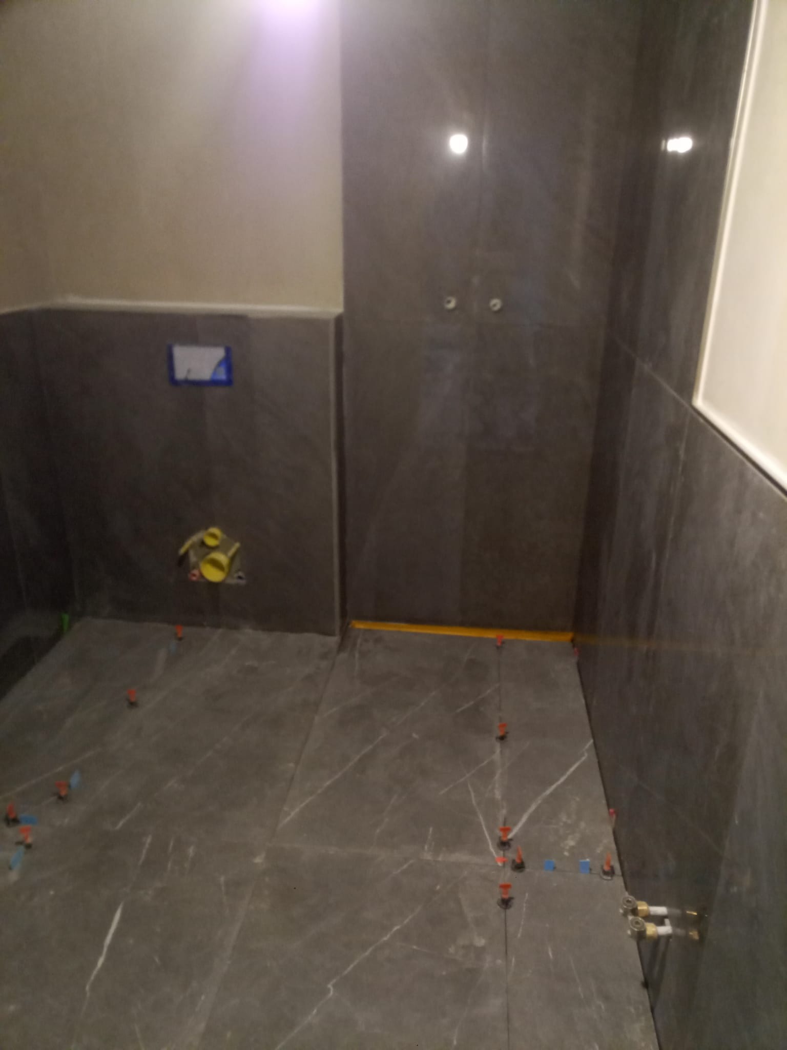 Układanie ciemnych płytek imitujących marmur w łazience, widoczne krzyżyki dystansowe i żółta rurka instalacyjna