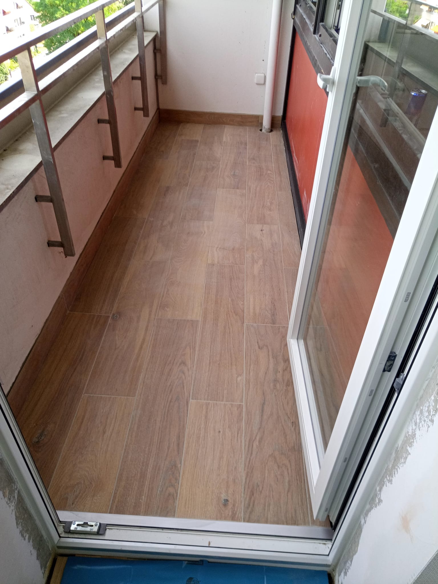 Wykończony balkon z drewnopodobnymi płytkami, białą stolarką okienną i metalową balustradą, widok z wnętrza mieszkania przez otwarte drzwi balkonowe.