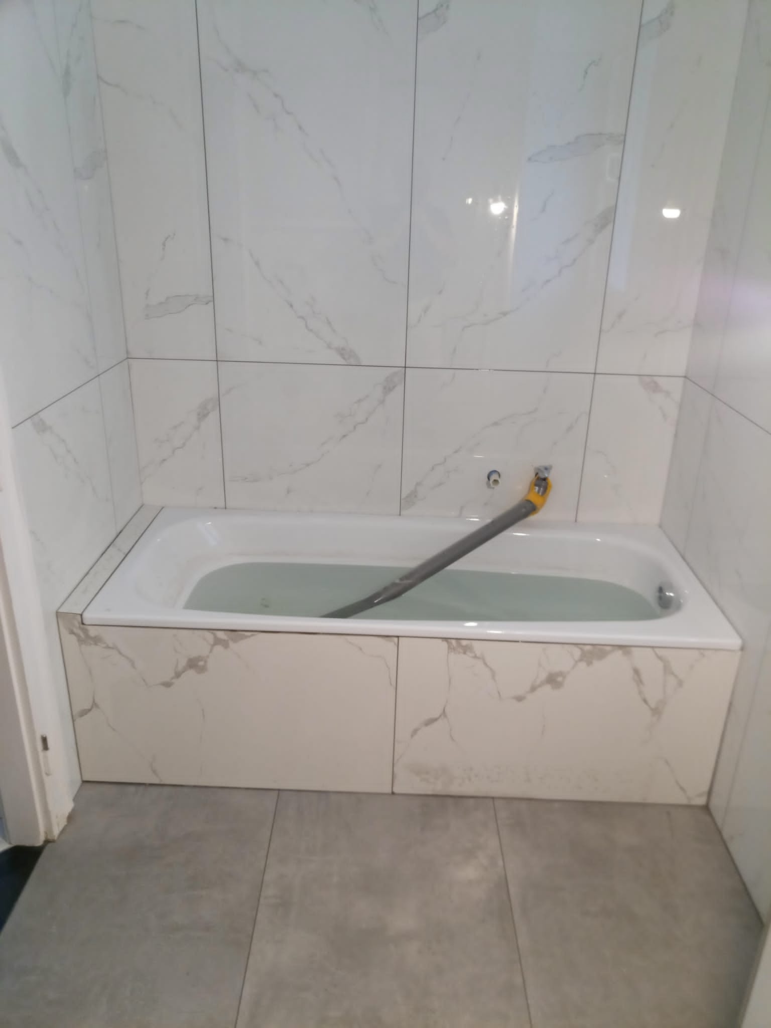 Wanna wypełniona wodą, obudowana płytkami imitującymi marmur, z szarym wężem odpływowym i białą glazurą na ścianach.