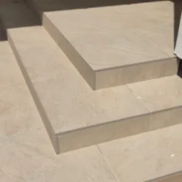 Zewnętrzne schody wejściowe wyłożone beżowymi płytkami ceramicznymi z widocznymi fugami, obok ściany z szorstkim tynkiem i czarną listwą przypodłogową, w tle widoczne akcesoria do czyszczenia.