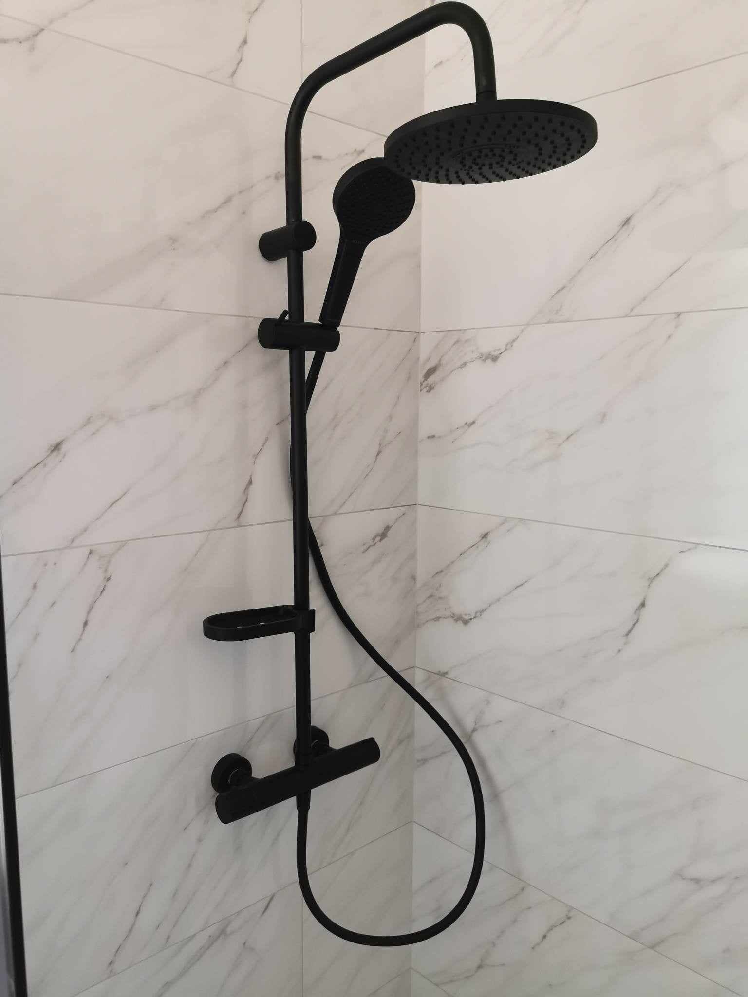 Czarny zestaw prysznicowy z deszczownicą i słuchawką, zamontowany na ścianie wyłożonej białymi płytkami imitującymi marmur.