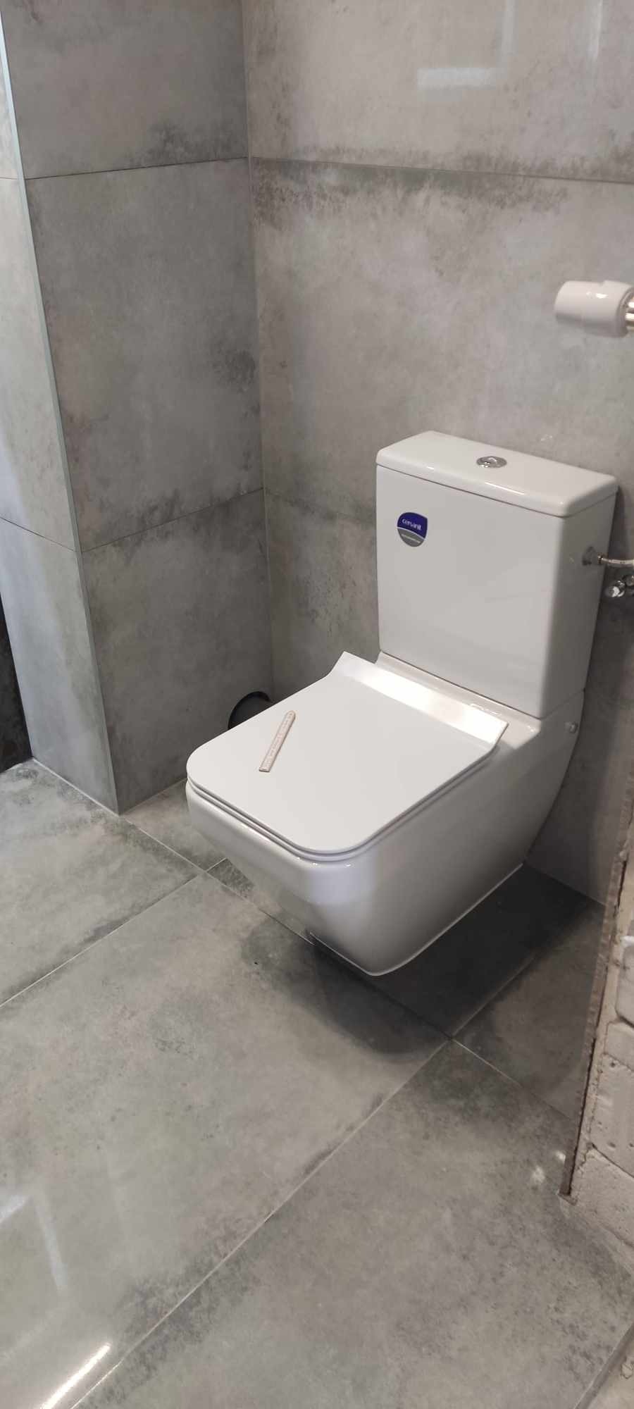 Nowoczesna toaleta wisząca z deską sedesową w kształcie kwadratu, zamontowana na szarych, betonopodobnych płytkach w łazience po remoncie, widoczny fragment ściany z płytkami i uchwyt na papier...