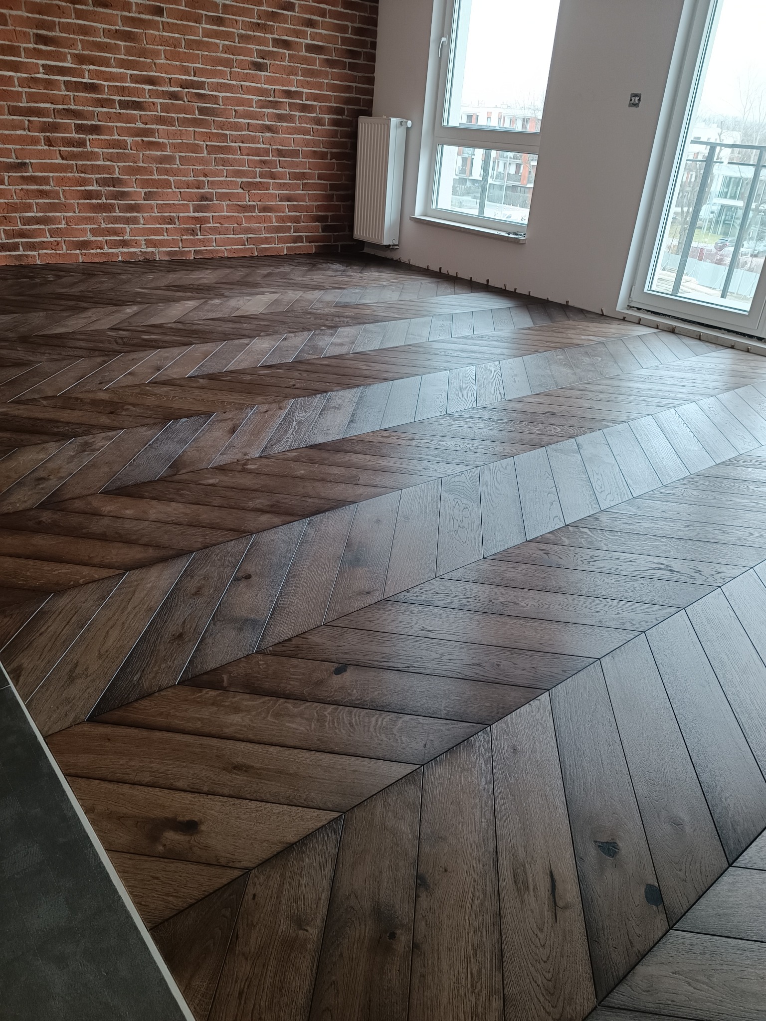 Ułożona jodełka z ciemnego drewna na podłodze w przestronnym pokoju z ceglaną ścianą i dużymi oknami balkonowymi.