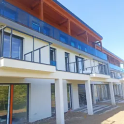 Nowoczesny budynek wielorodzinny z balkonami z niebieskimi szklanymi balustradami, świeżo po ociepleniu elewacji, widoczne przygotowane podłoże pod taras.