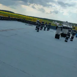 Płaski dach pokryty nową, szarą membraną bitumiczną z przygotowanymi kominami wentylacyjnymi, widok na oddalony krajobraz z polami i domami.