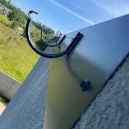 Czarne, metalowe uchwyty rynnowe zamocowane do ciemnej, gładkiej powierzchni dachu, widok z dołu na błękitne niebo i zieleń w tle.