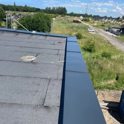 DachSystem Łukasz Ślęzak - Widok z góry na fragment dachu pokrytego papą, z zamontowanymi, szarymi obróbkami blacharskimi przy krawędzi, zwinięty kabel i narzędzia na powierzchni, w tle krajobraz z drzewami i domami.