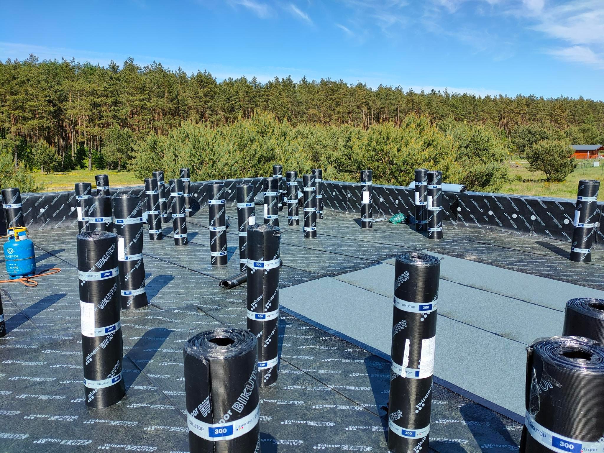 Przygotowanie do krycia dachu papą termozgrzewalną Bikutop 300, widoczne rolki papy i butla gazowa, w tle las.
