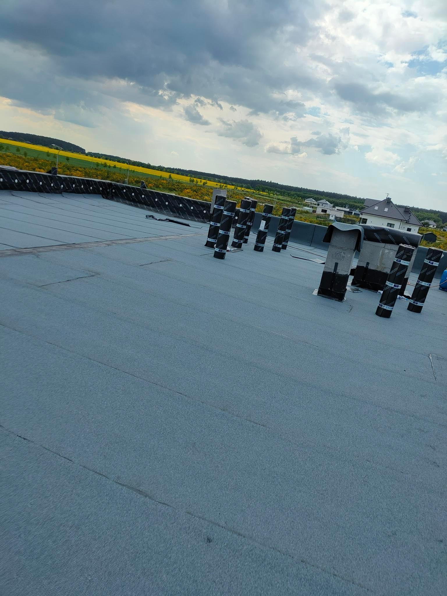 Płaski dach pokryty nową, szarą membraną bitumiczną z przygotowanymi kominami wentylacyjnymi, widok na oddalony krajobraz z polami i domami.