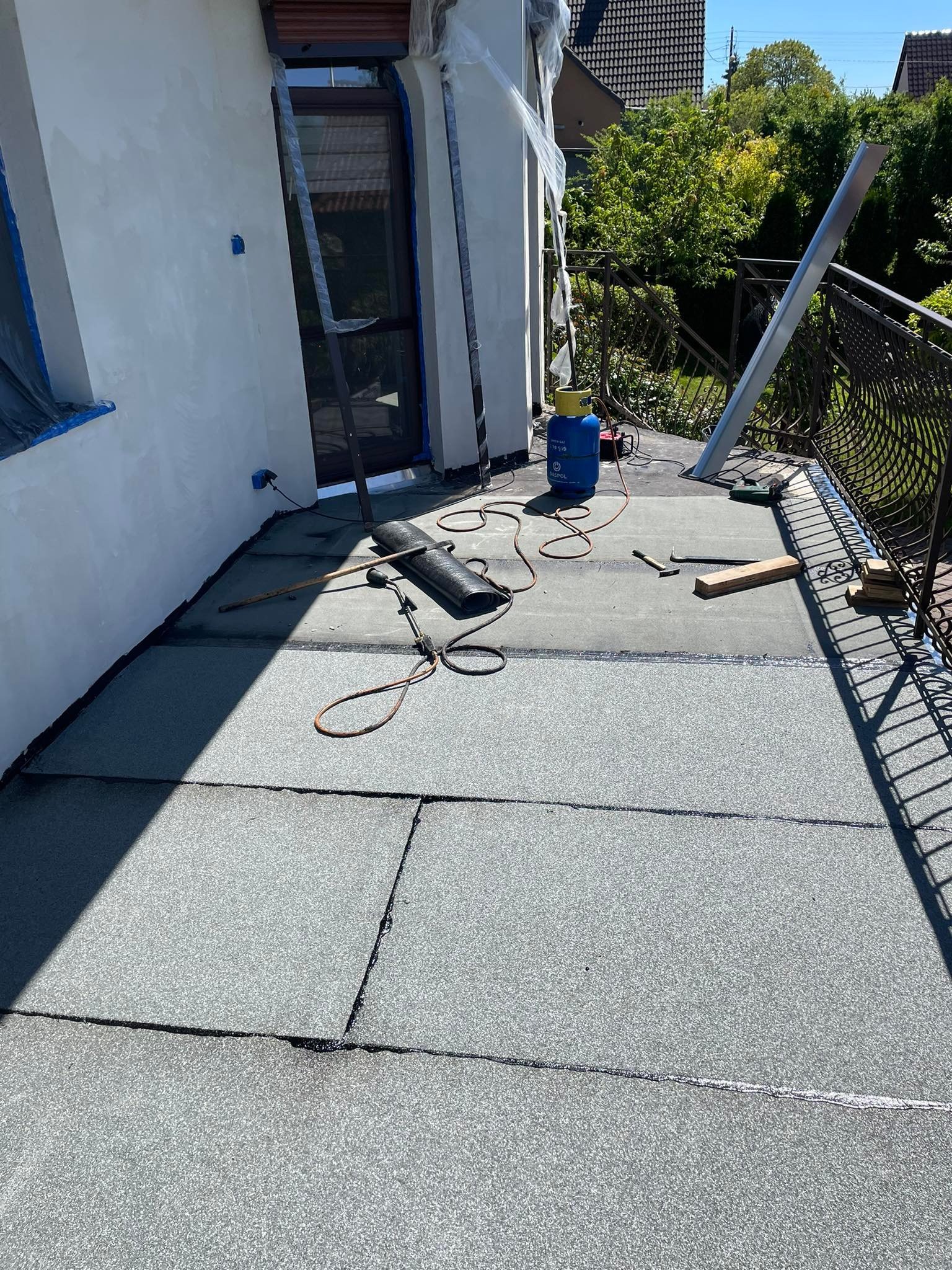 Montaż papy termozgrzewalnej na balkonie z użyciem palnika, widoczna butla gazowa, narzędzia dekarskie i zabezpieczone okno folią malarską.